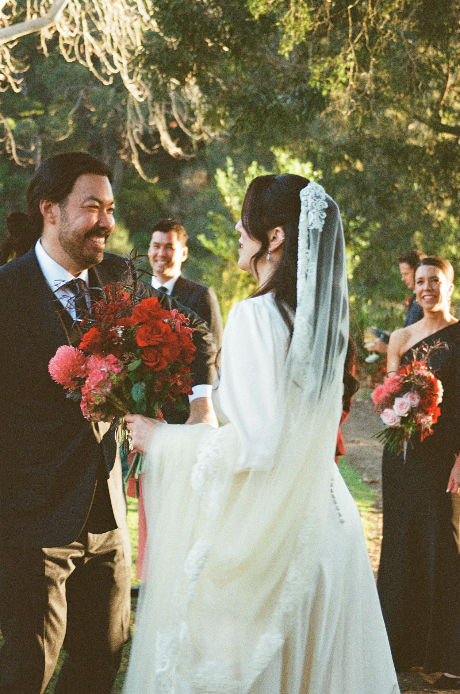 A romantic outdoor wedding ceremony with beautiful red floral bouquets and a trailing bridal veil in golden sunlight.