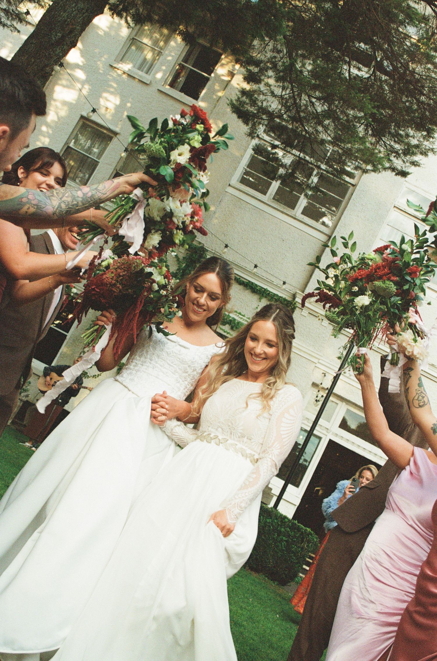 Two brides in white gowns walk through a floral arch held by wedding guests outdoors.