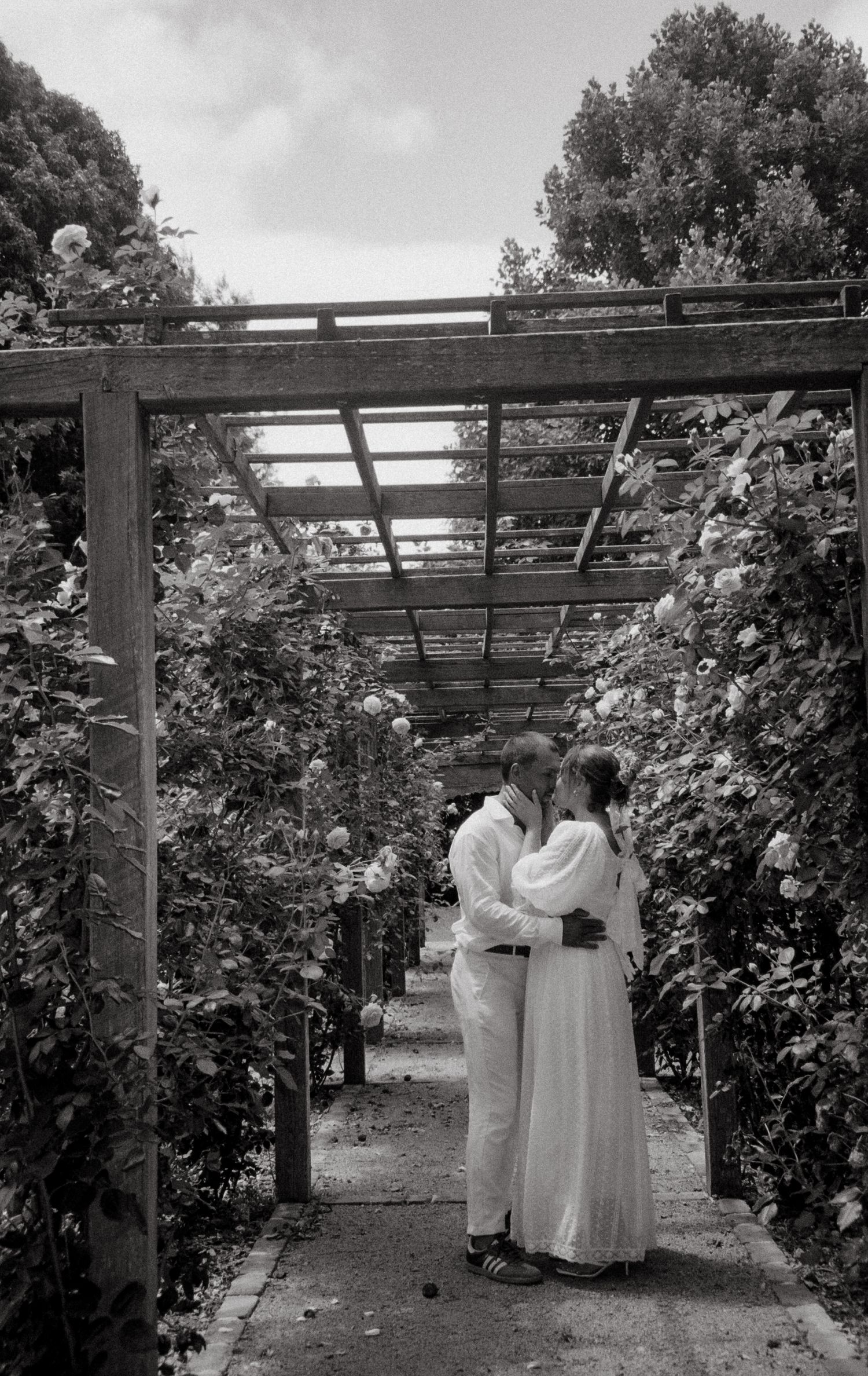 A romantic black and white photo of a couple embracing under a garden pergola surrounded by climbing vines.