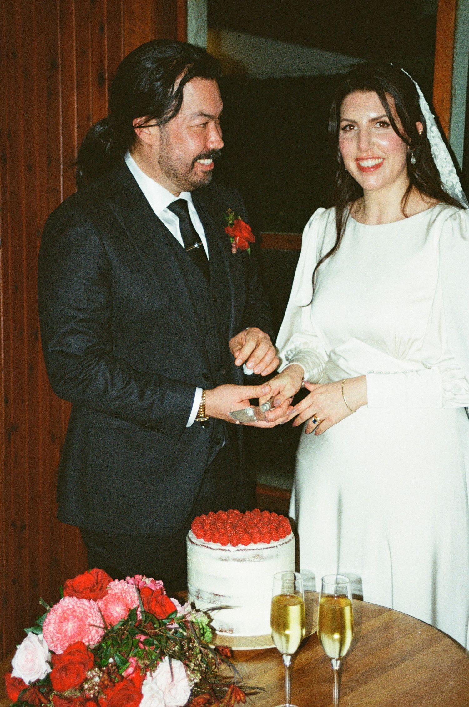 A couple exchanges rings during their wedding ceremony with champagne and a cake decorated with red flowers.