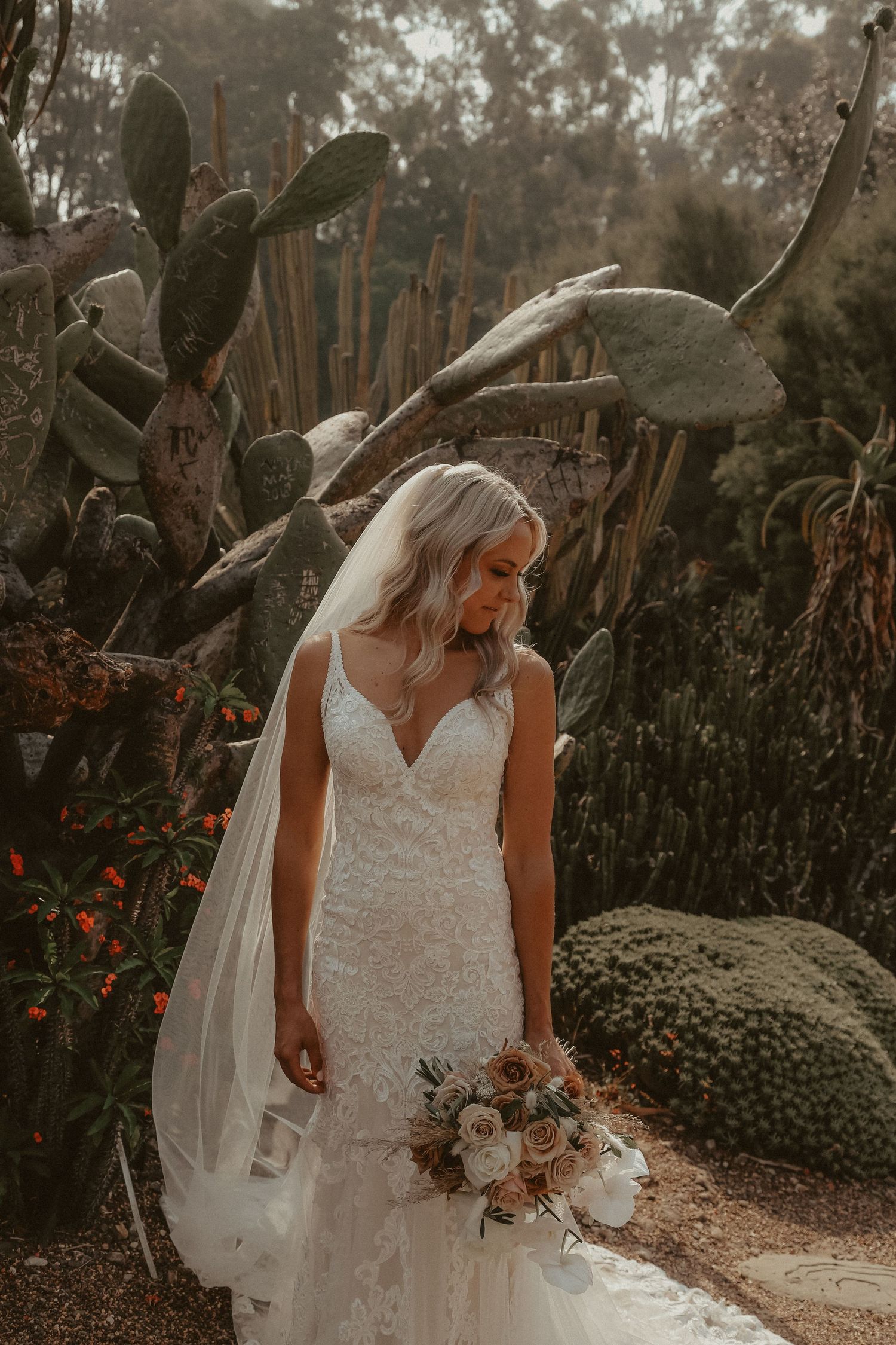 A bride in a white lace wedding dress poses in a moody outdoor garden setting with flowering plants at sunset.
