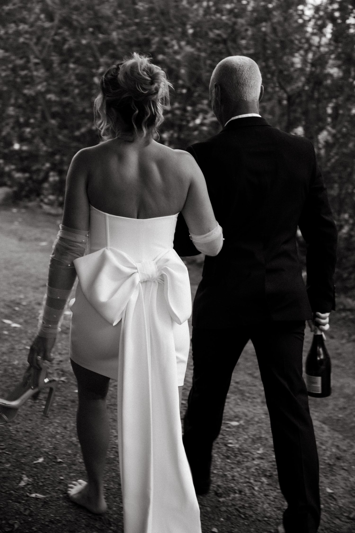 A wedding couple walks along a garden path in black and white, with the bride's dress featuring a large bow.