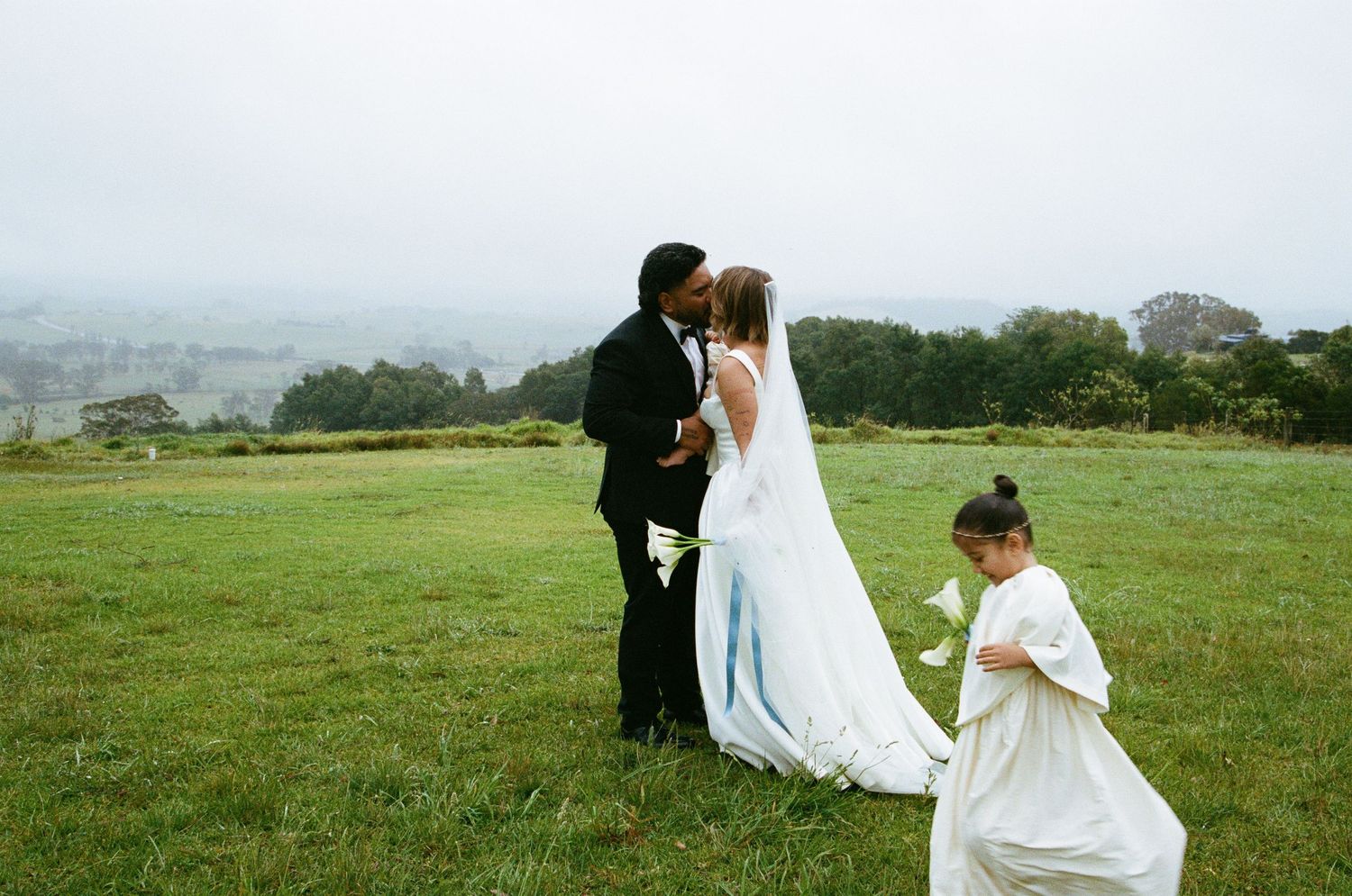 A wedding couple shares a moment on a misty hilltop while an attendant adjusts the bride's flowing white dress train.