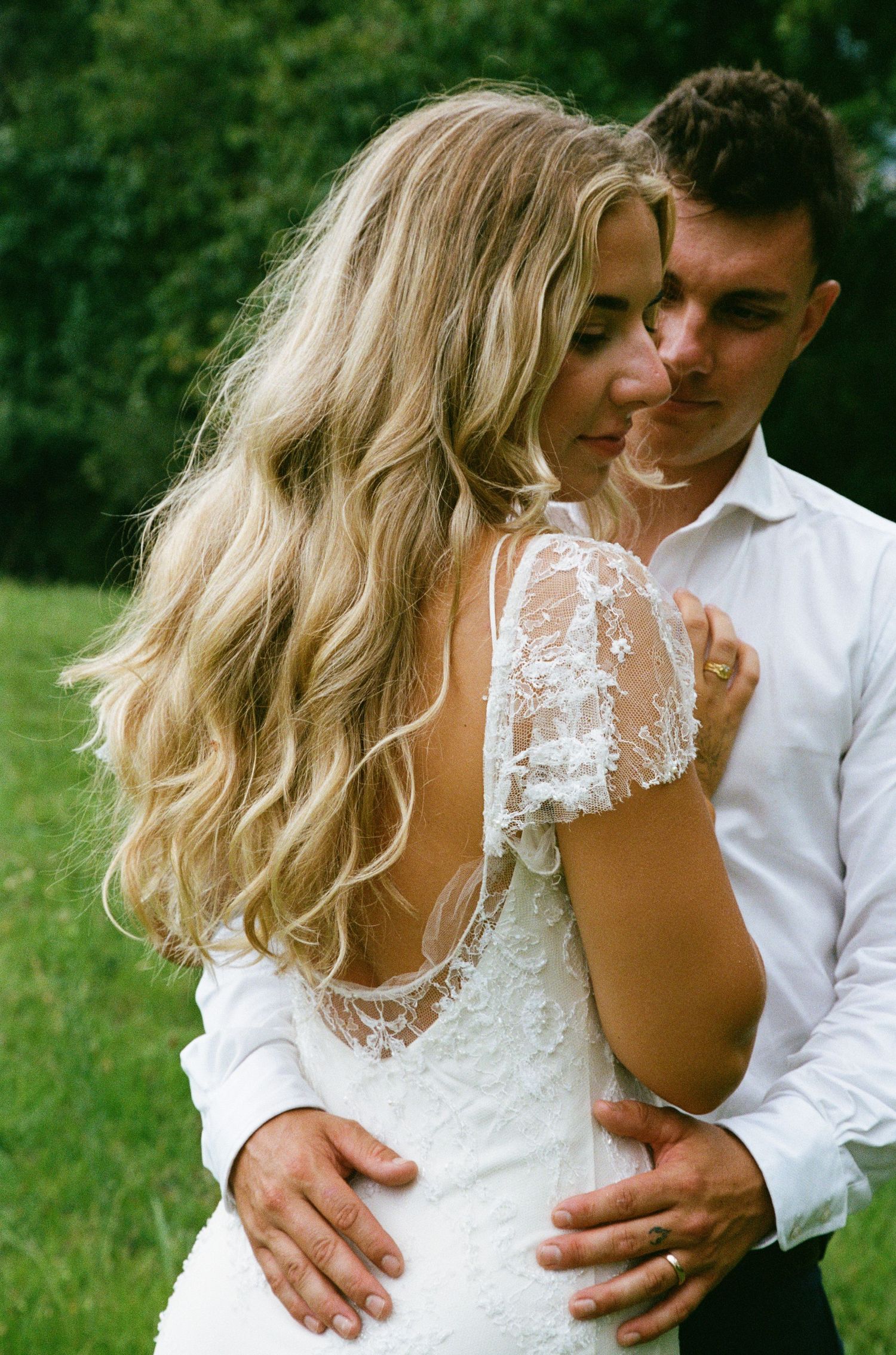 A romantic wedding couple embraces outdoors in a natural setting with lush greenery in the background.