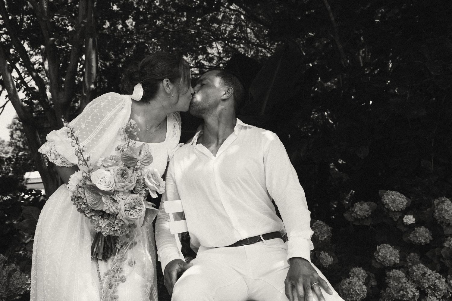 A romantic black and white outdoor wedding photo showing a tender moment with flowers and trees in the background.