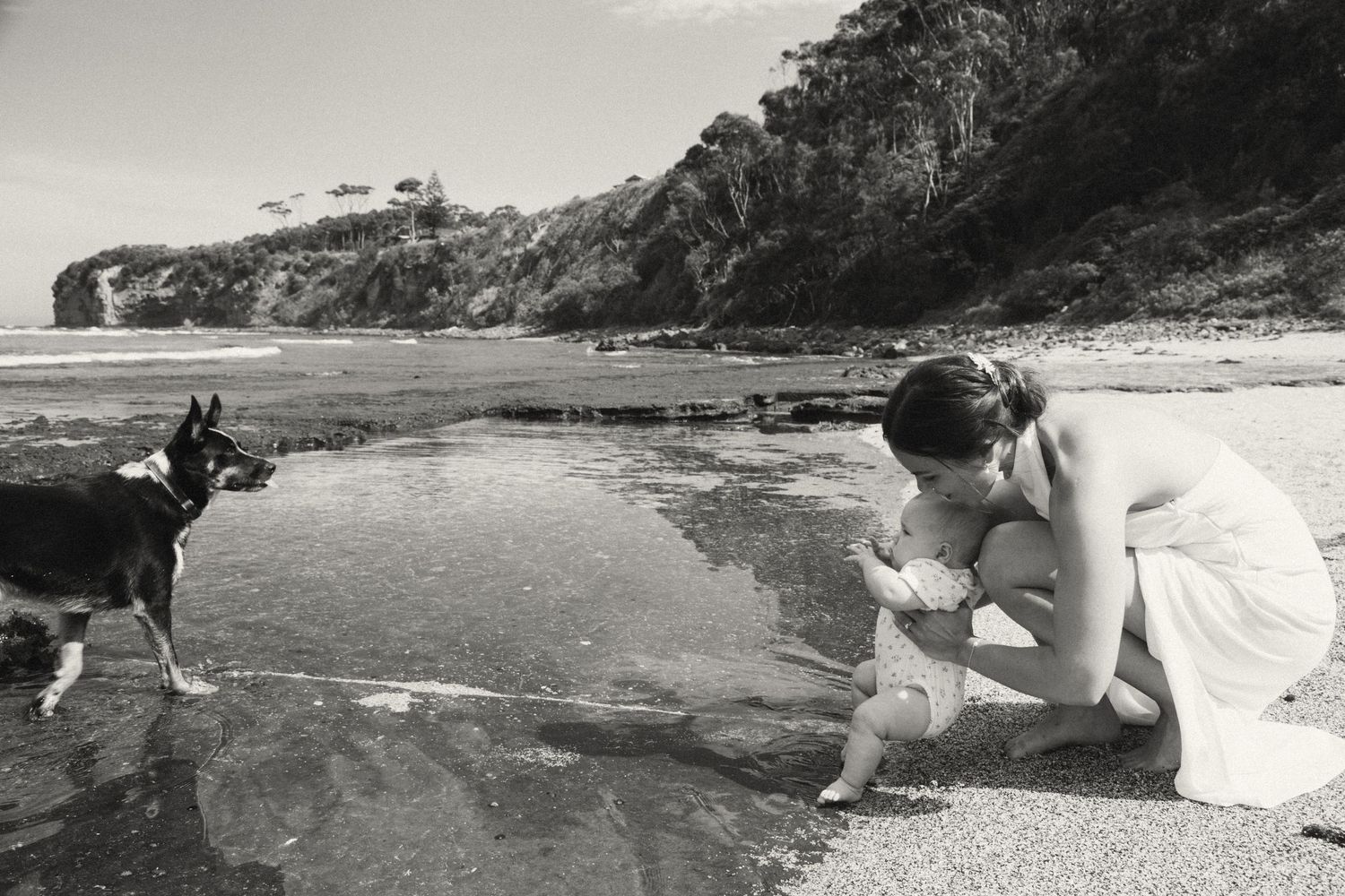 Black and white photo of someone in a white dress crouching by a tide pool while a dog stands nearby on the beach.