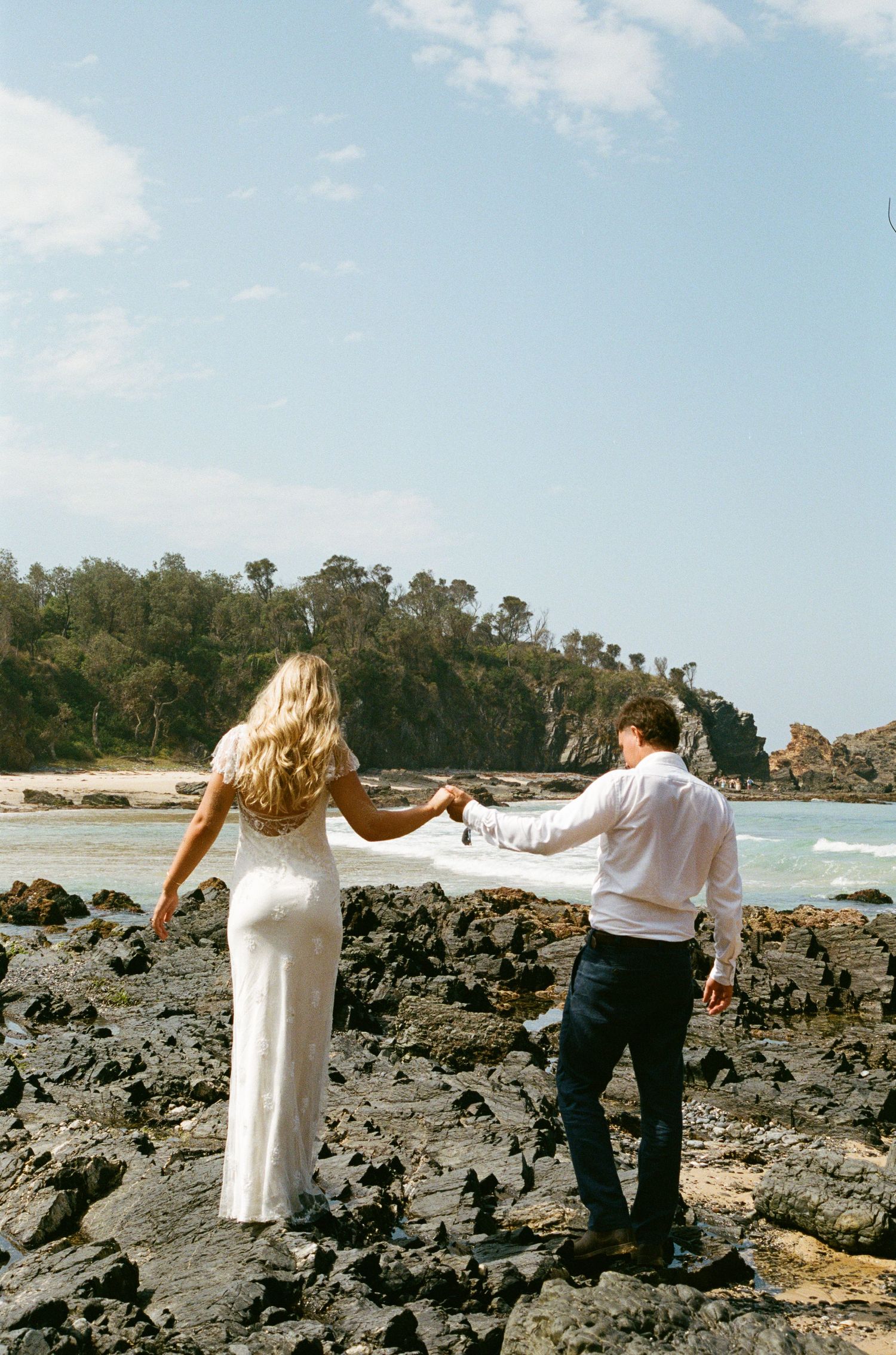 A couple walks hand in hand across rocky coastal terrain with ocean waves in the background.