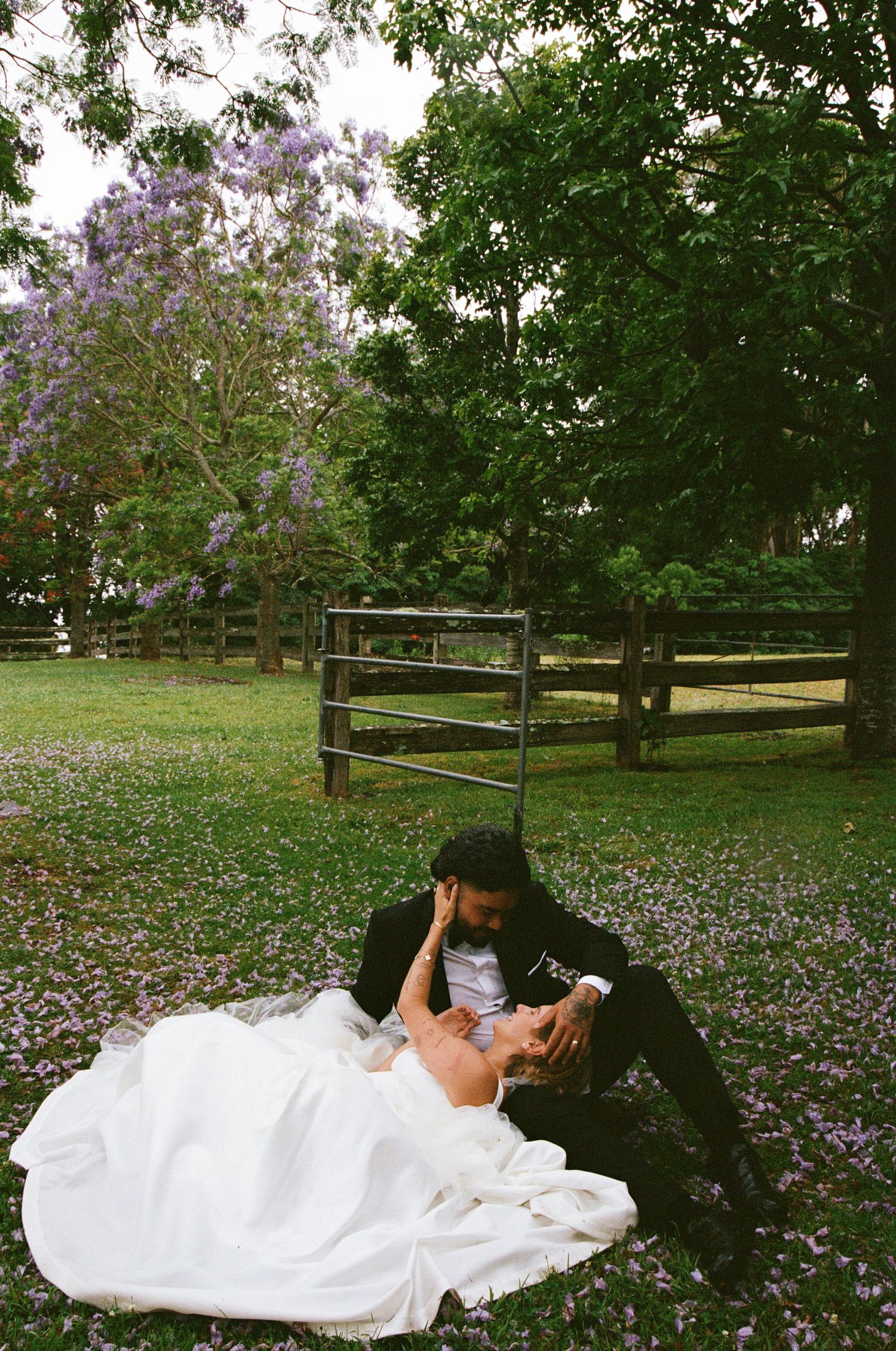 Newlyweds share an intimate moment on grass beneath blooming lilac trees.