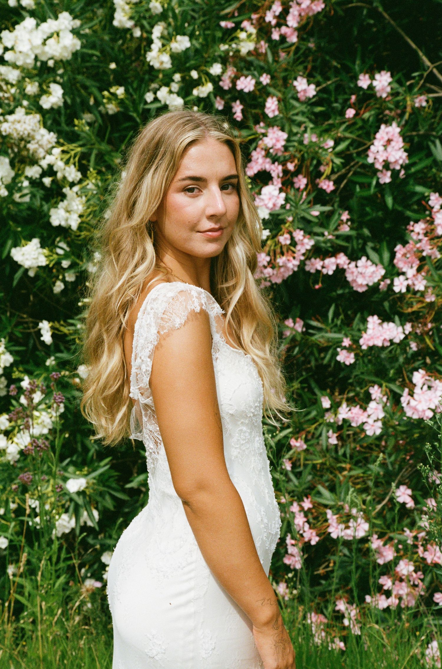Bride in a lace wedding dress poses against a backdrop of pink and white flowers in a garden setting.