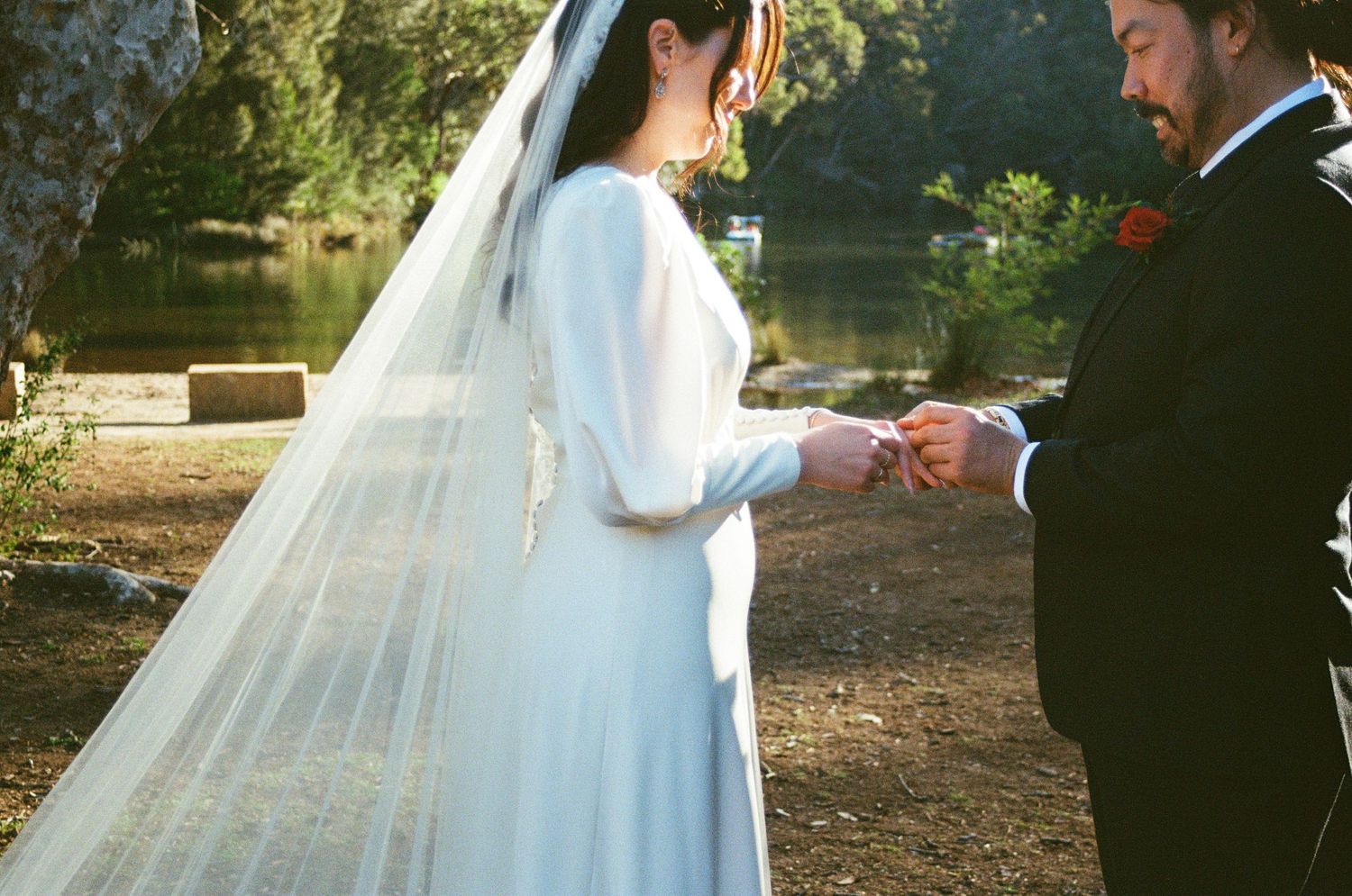 A couple exchanges rings during an outdoor wedding ceremony by a lake, with sunlight filtering through trees.