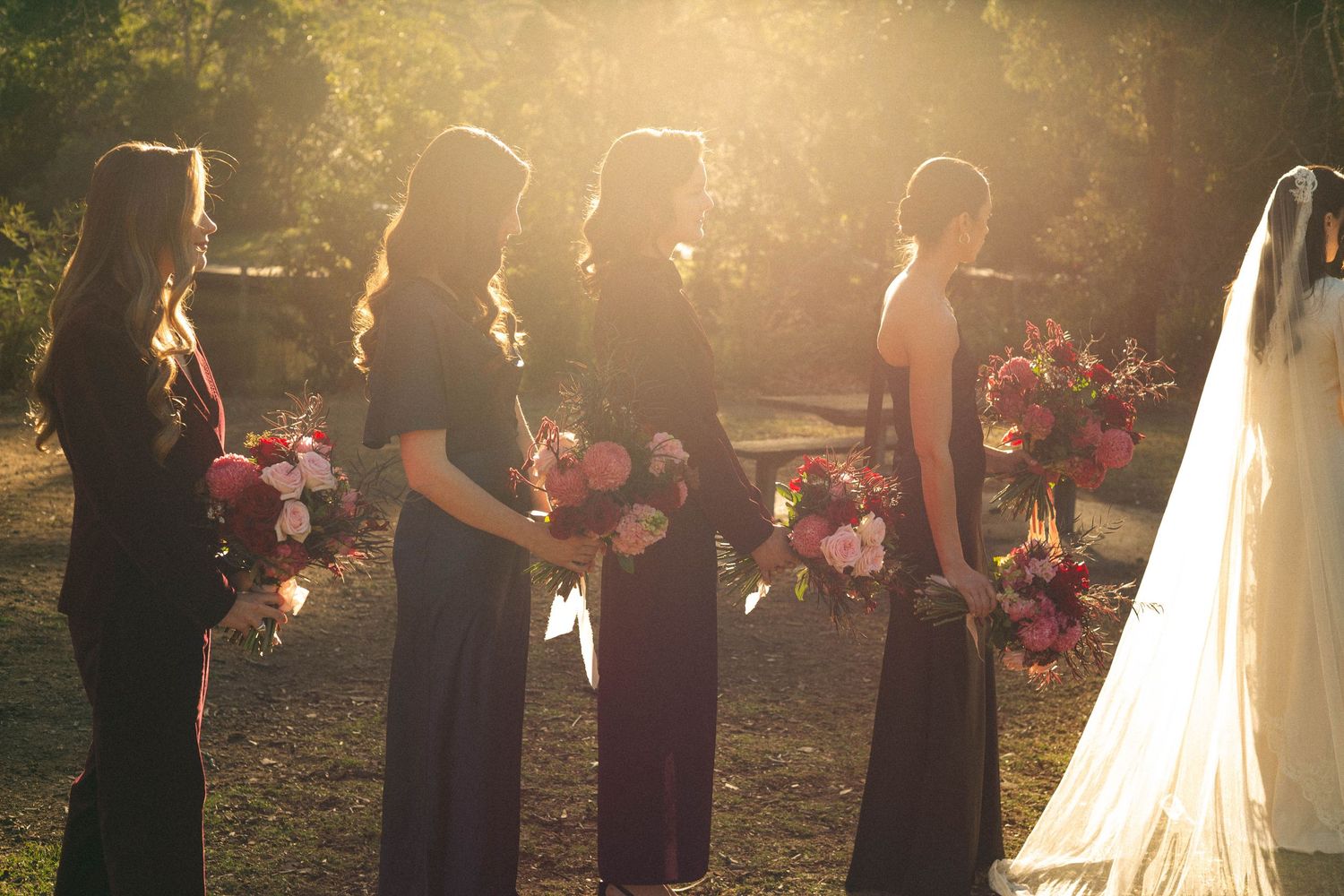 Bridesmaids in dark dresses stand in a row during a sunset wedding ceremony.