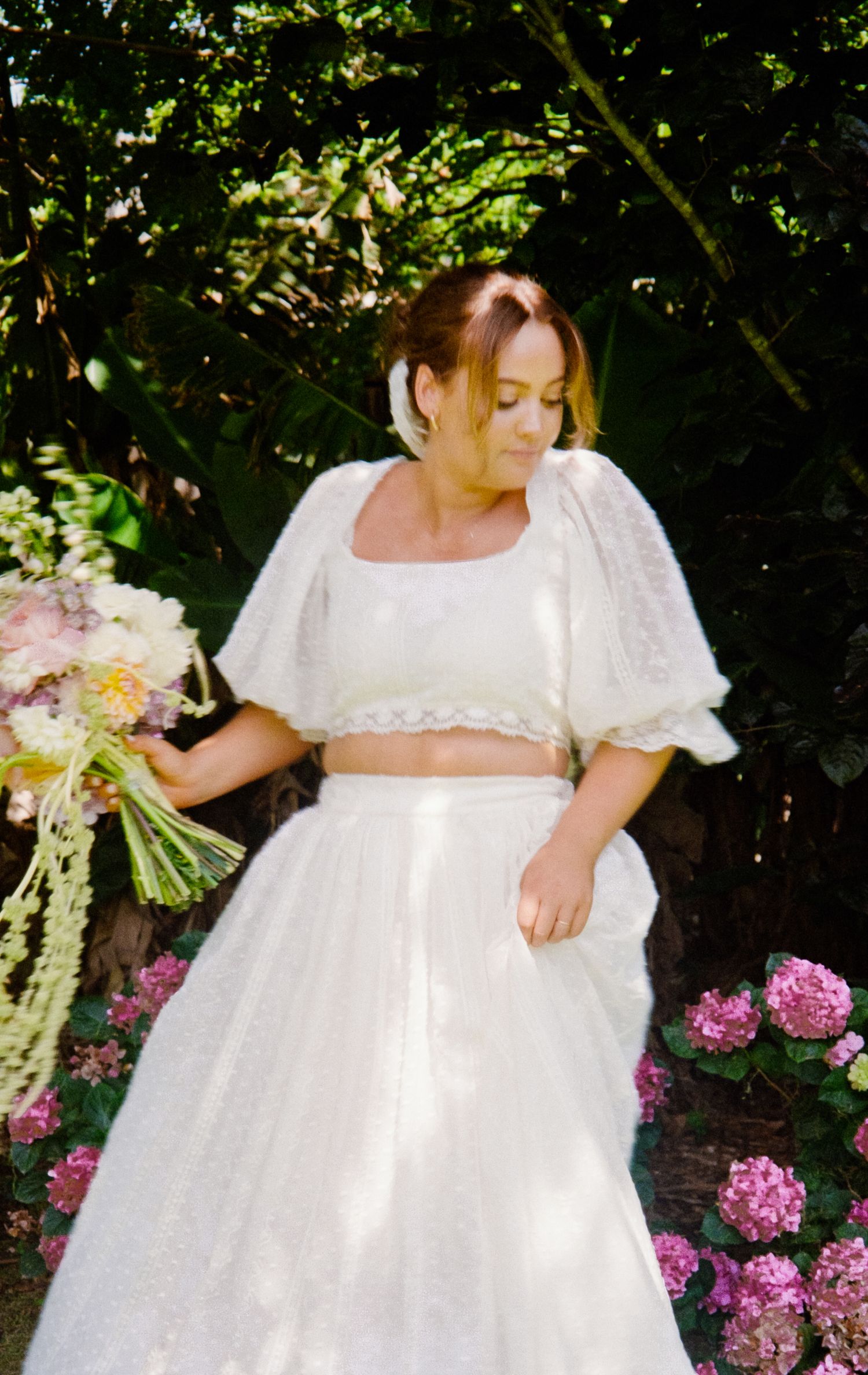 Two-piece bridal gown with cropped top and full skirt showcased against pink hydrangeas in a garden.