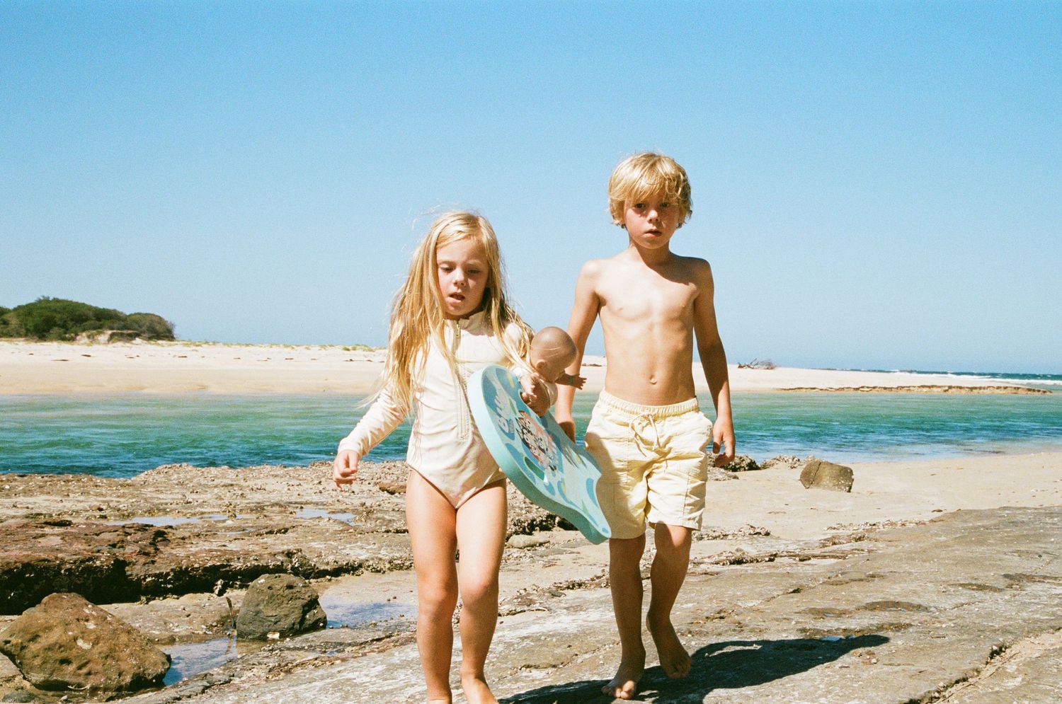 Two children in swimsuits walk along a rocky beach shore on a sunny summer day with blue ocean waters in the background.