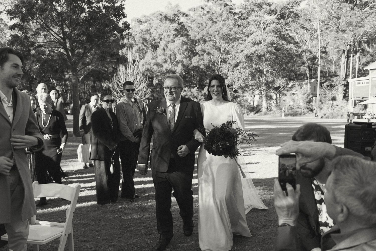 A vintage black and white photo of a bride walking down an outdoor aisle with guests looking on.
