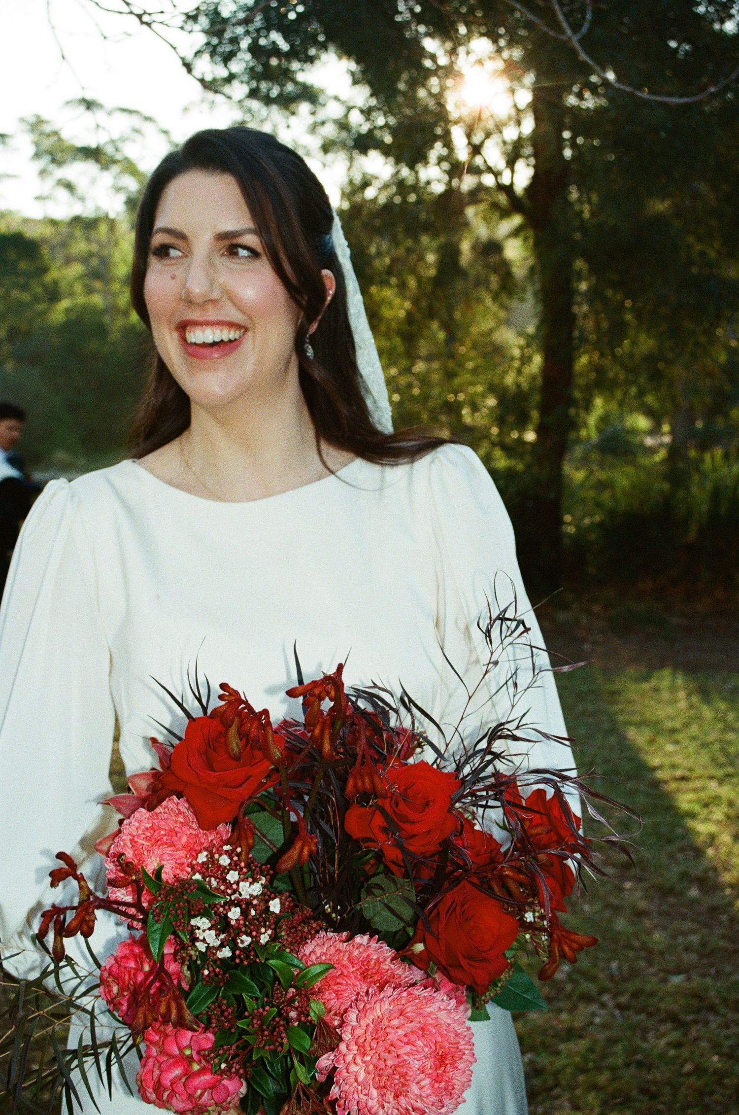A bride wearing a classic white dress holds a vibrant bouquet of red and pink flowers against a sunlit outdoor backdrop.
