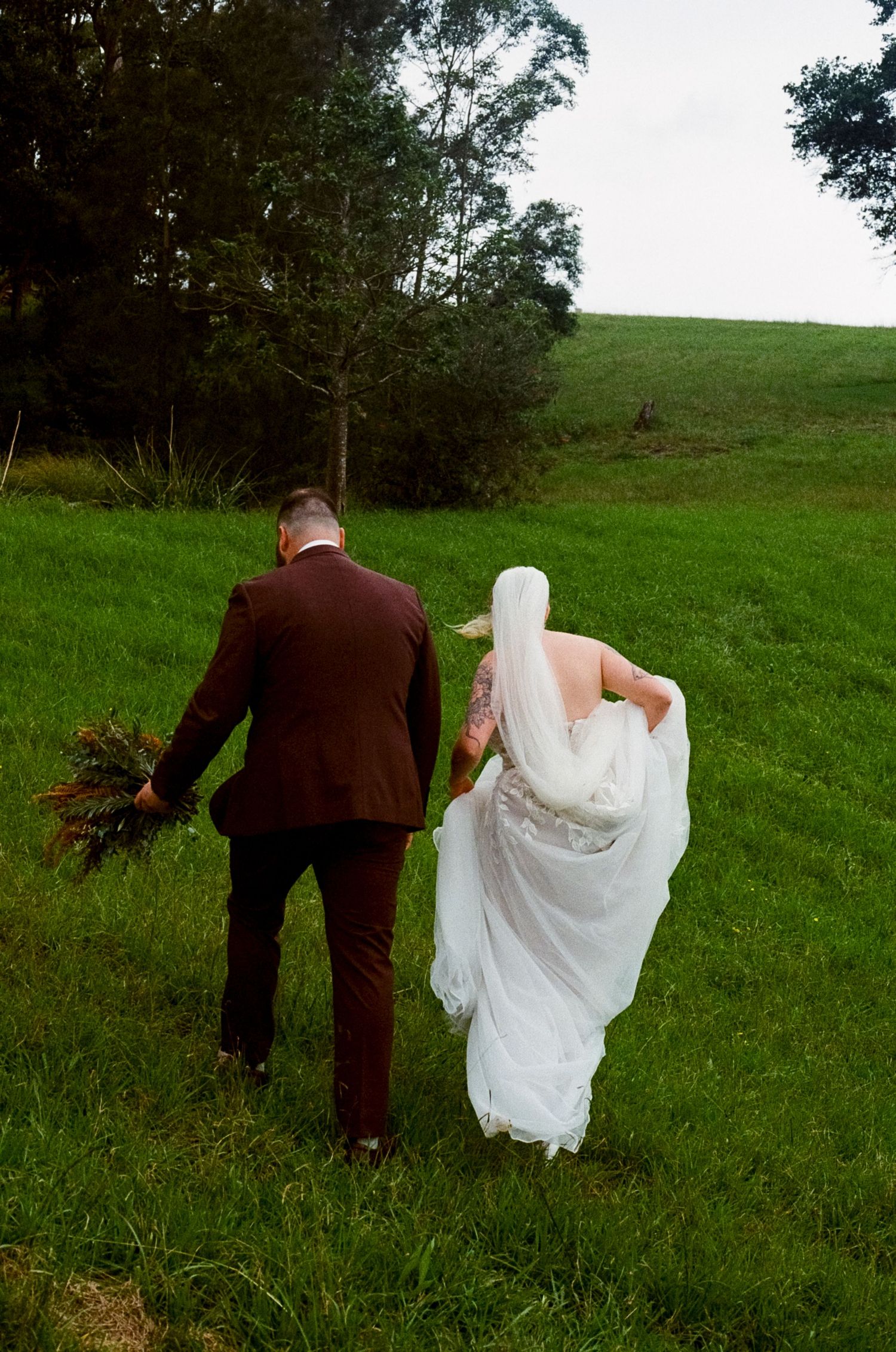 A couple in wedding attire walks together across a green hillside in a romantic outdoor setting.