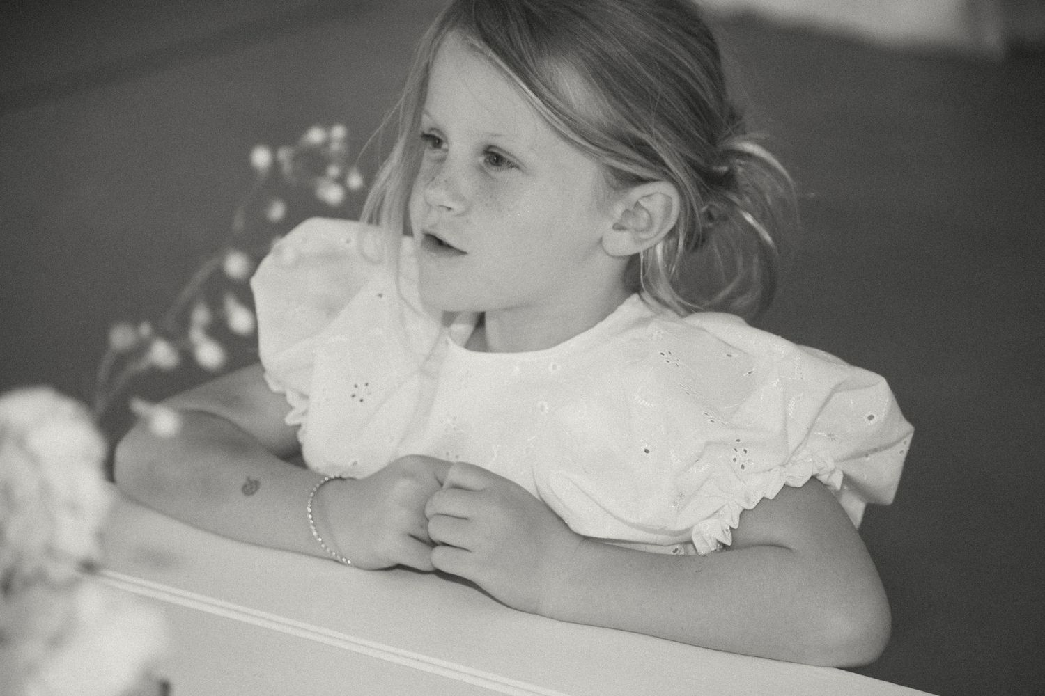 A black and white portrait of a young child in a white dress with lace collar sitting at a table.