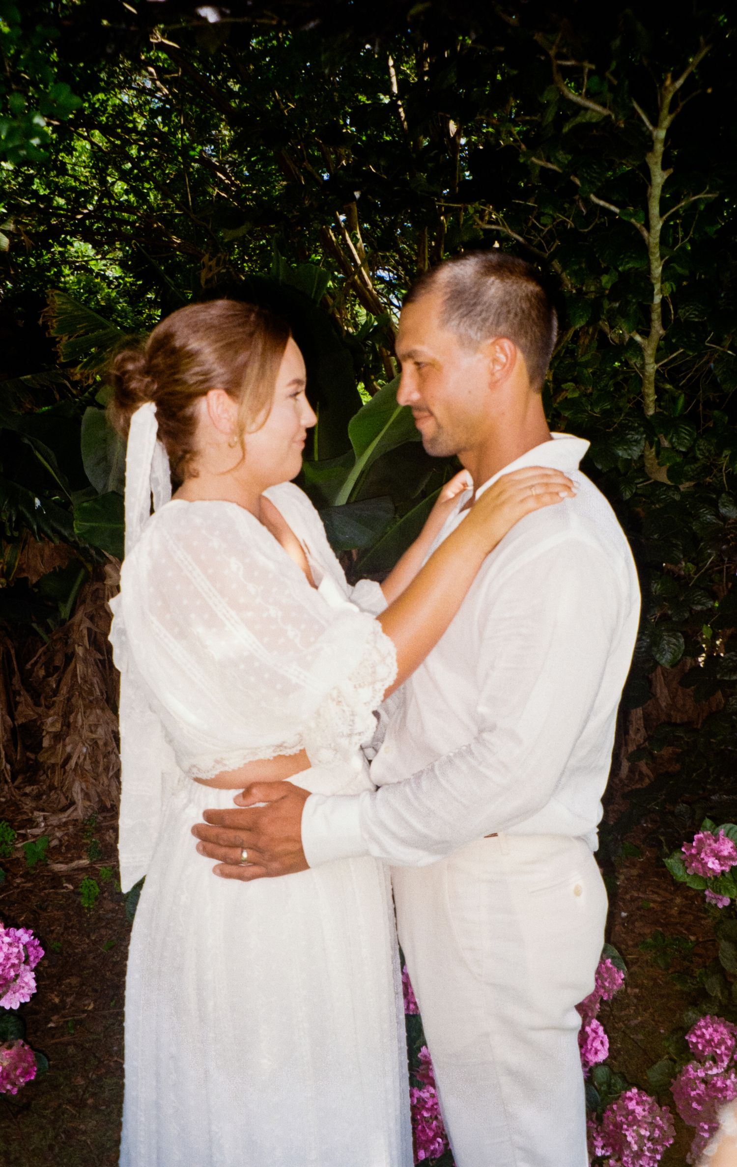 A couple in white outfits dances together in a garden with pink flowers blooming in the background.