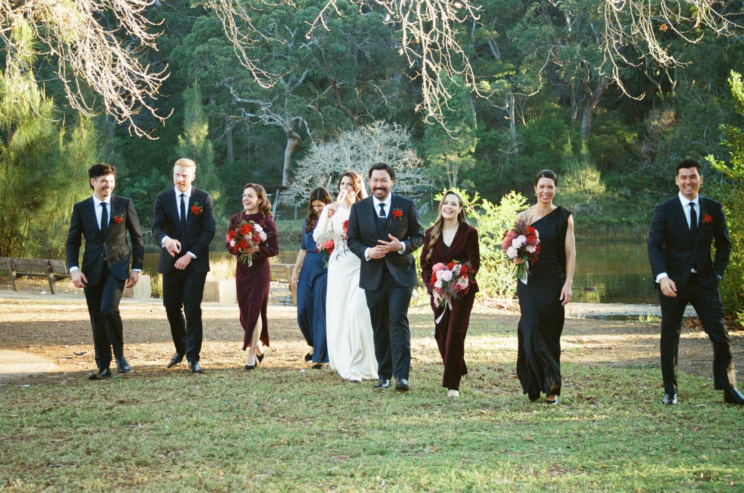 Wedding party lines up in a scenic outdoor garden setting for group photos in natural sunlight.