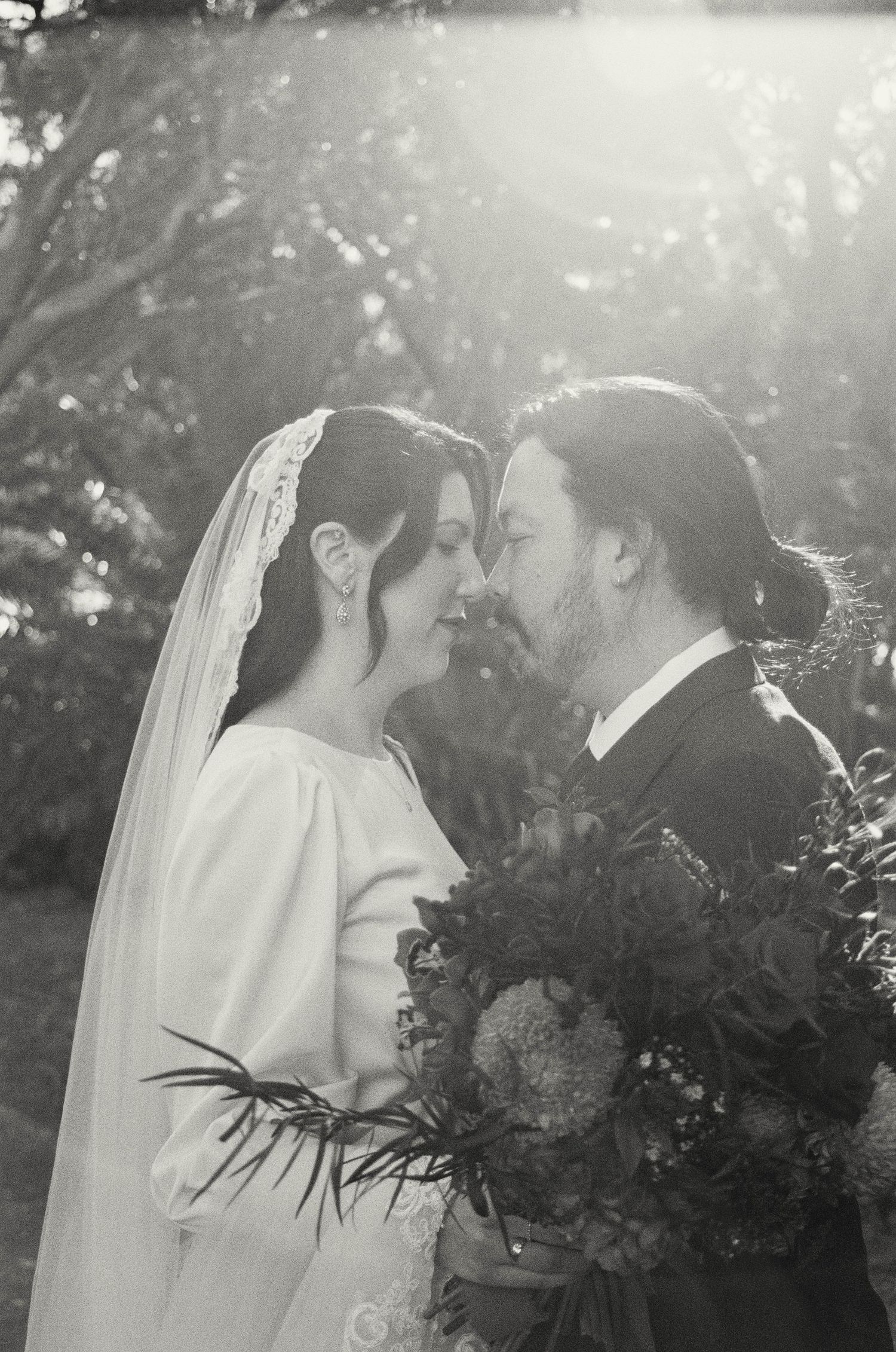 A romantic wedding scene in black and white with soft sunlight filtering through trees.