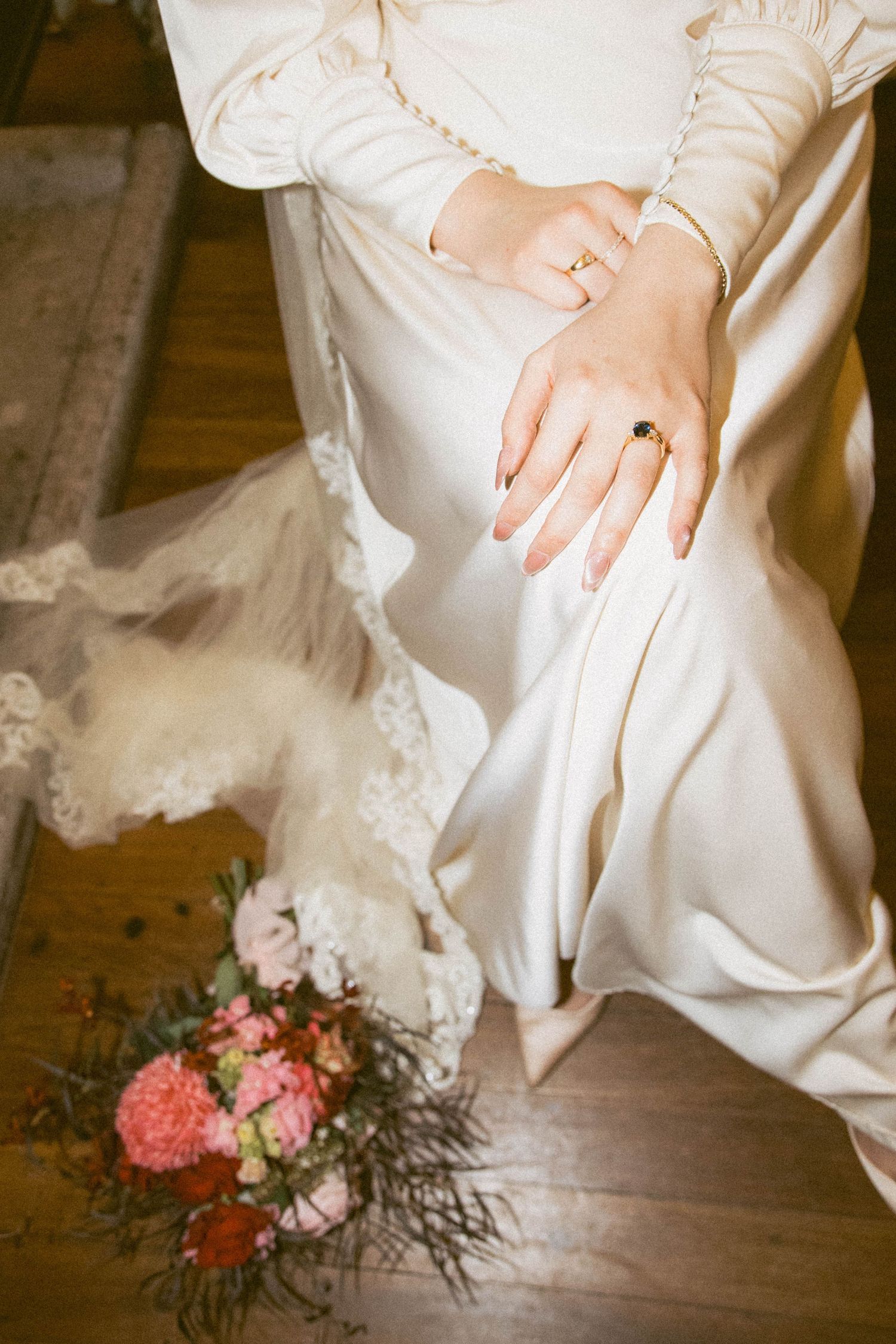 Close-up of bride's hands with wedding rings and white dress, pink floral bouquet on wooden floor.