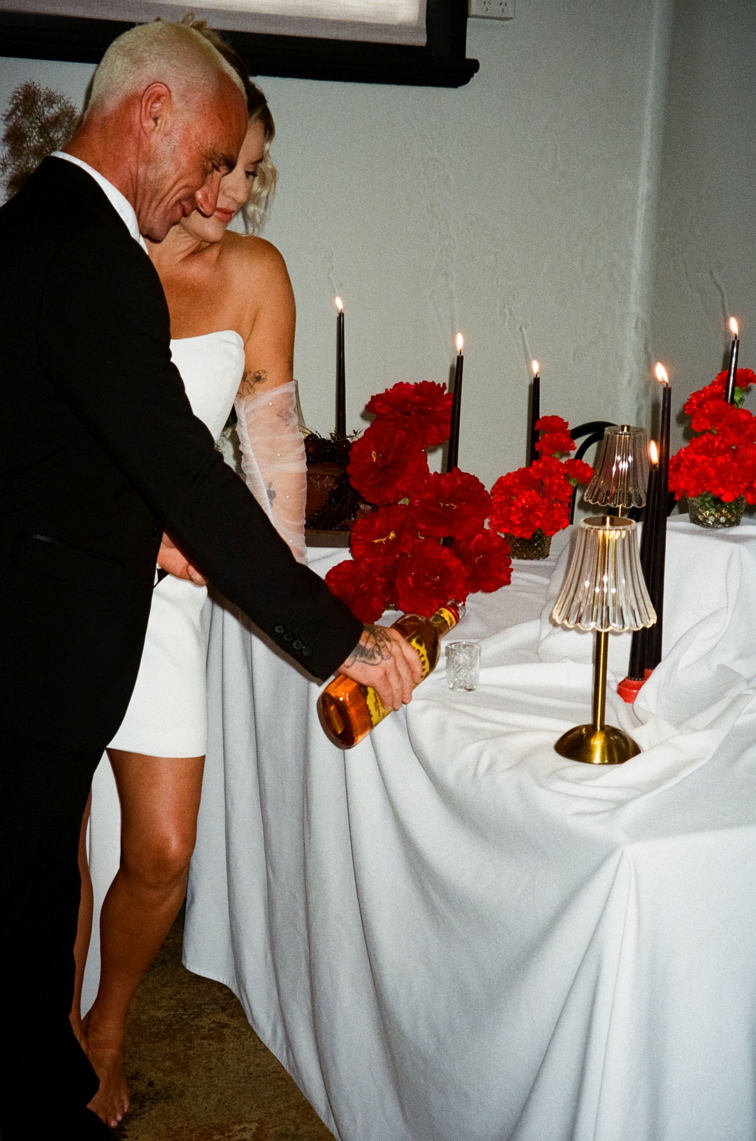 Person in formal attire arranges elegant table settings with white linens, red roses, and lit candles at a banquet event.