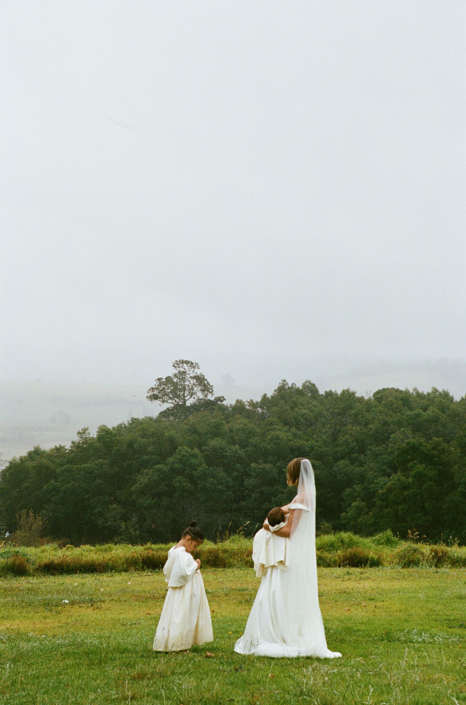 A bride and two young attendants in white dresses stand in a misty field.