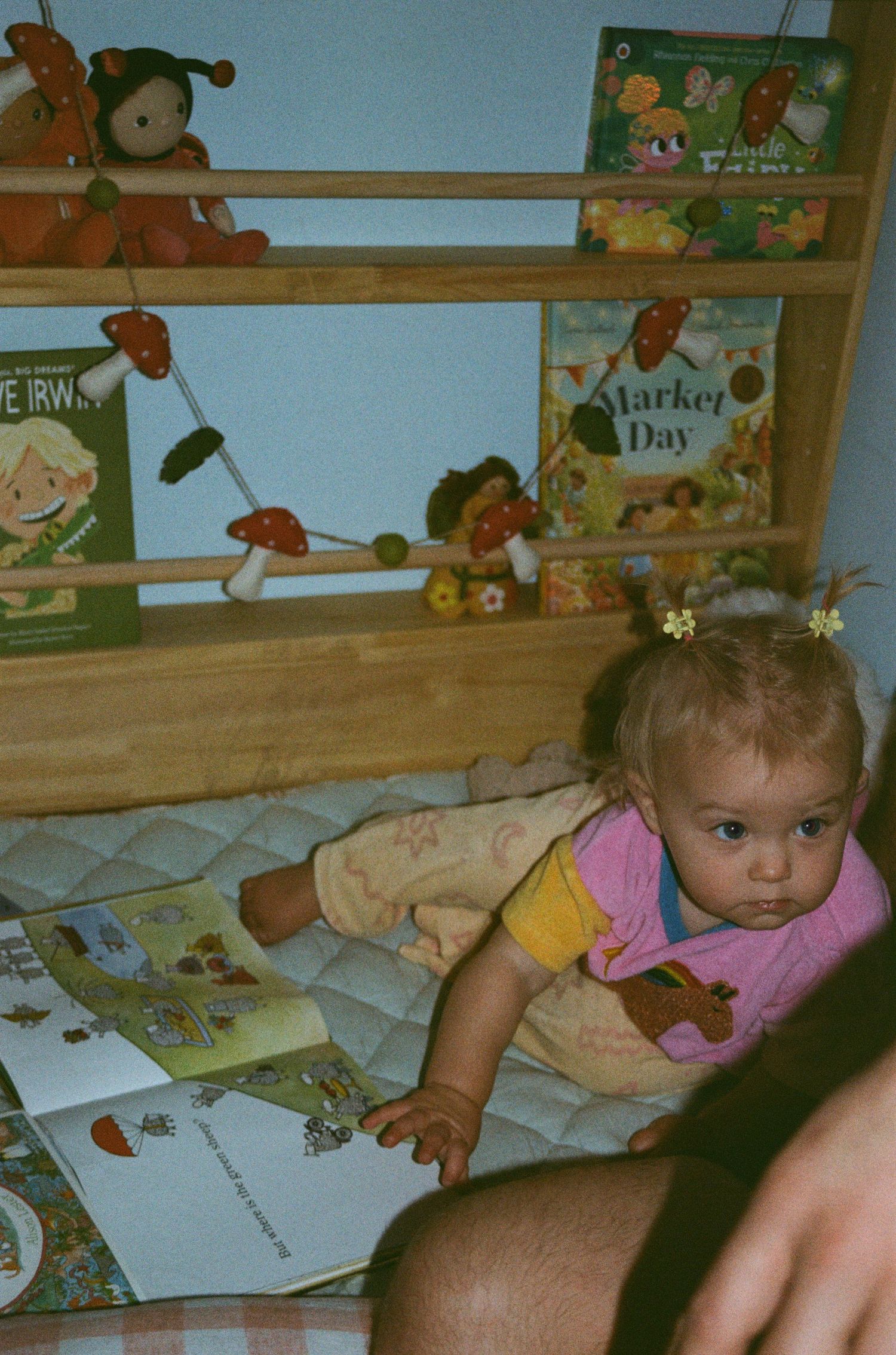A young child lies on the floor looking at picture books with a wooden bookshelf displaying children's stories in the background.
