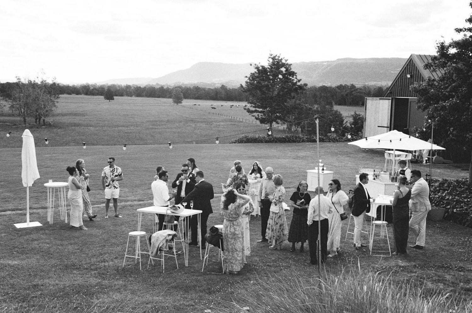 A vintage black and white photograph shows a group of people gathered at an outdoor social event with tables and chairs.
