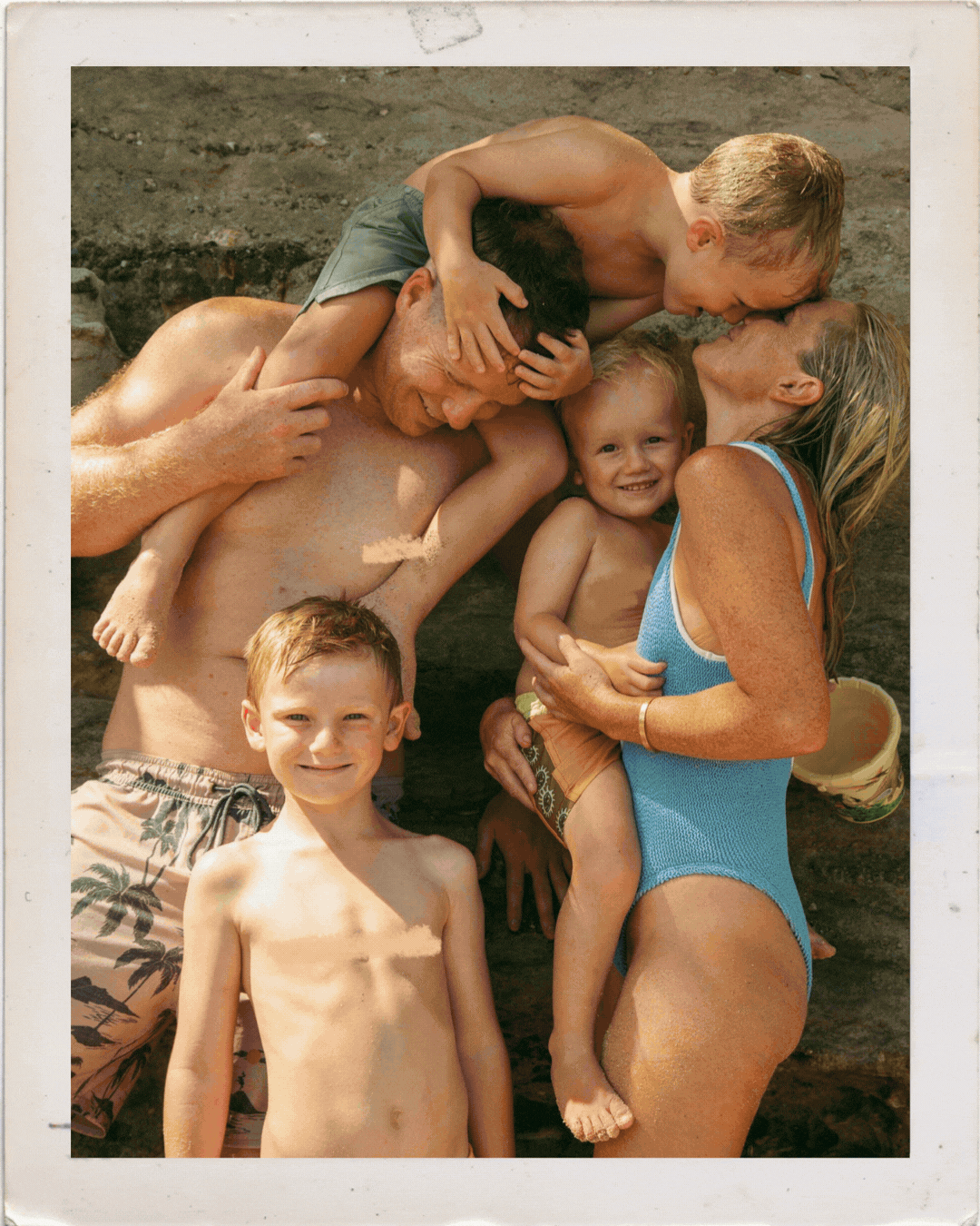 Family photo at the beach with parents and three young children hugging and smiling together outdoors.