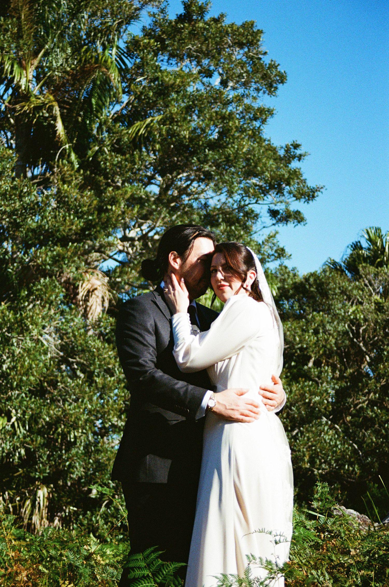 Wedding couple embraces amidst green garden foliage beneath sunny blue sky.