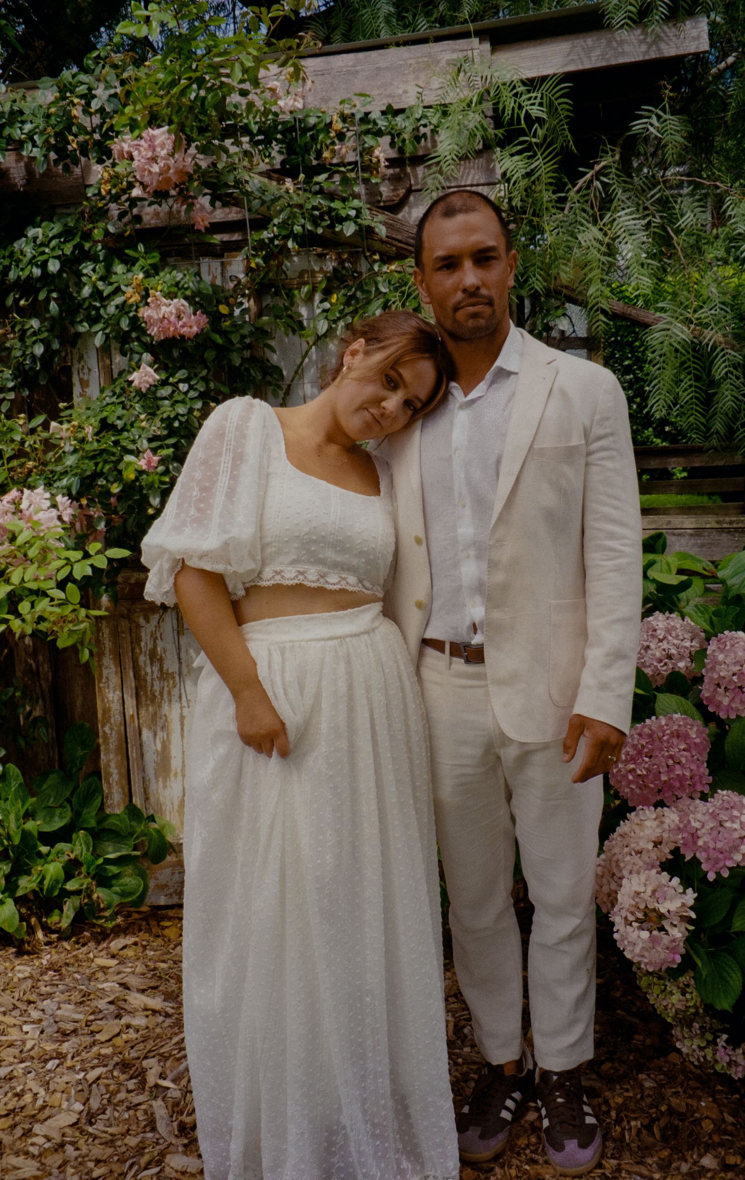 A romantic couple in white vintage clothing poses together in a garden with blooming flowers.