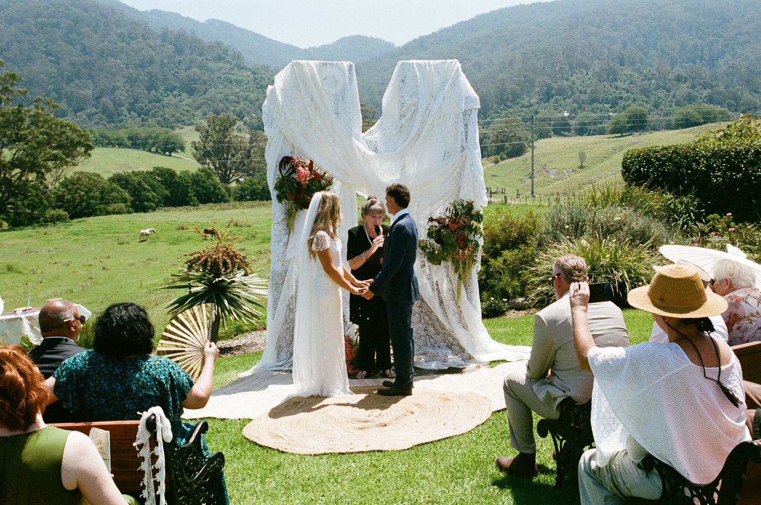 Wedding guests gather under white fabric draping with rolling hills and mountains in the background.
