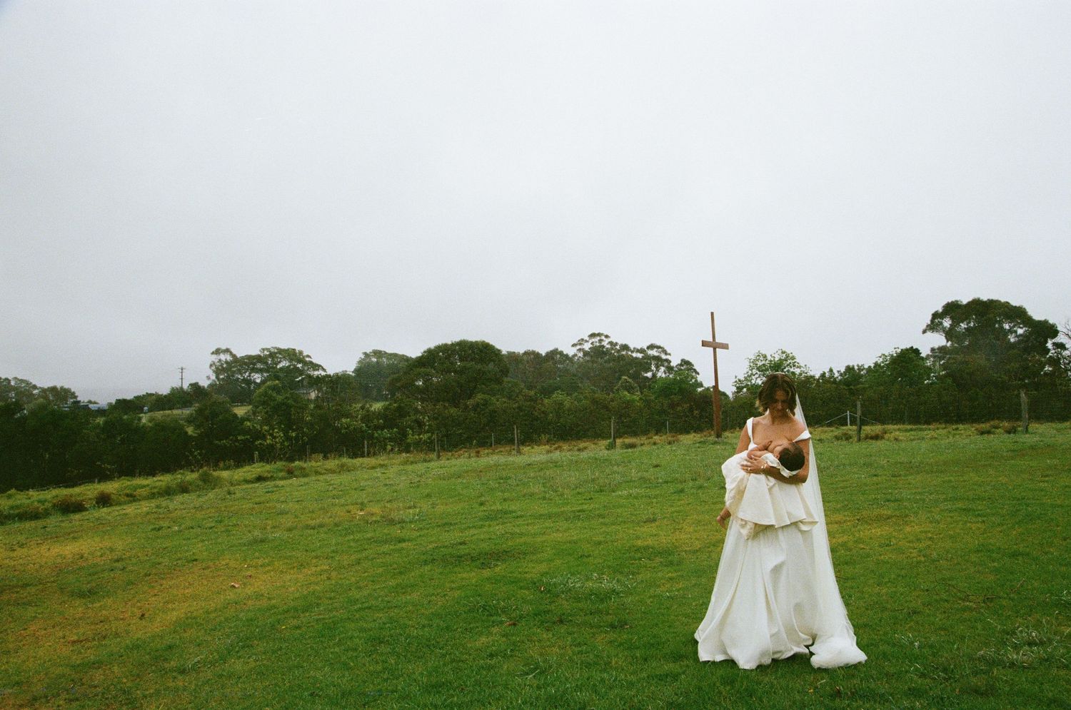 A couple embraces on a grassy hilltop with a cross visible in the background.