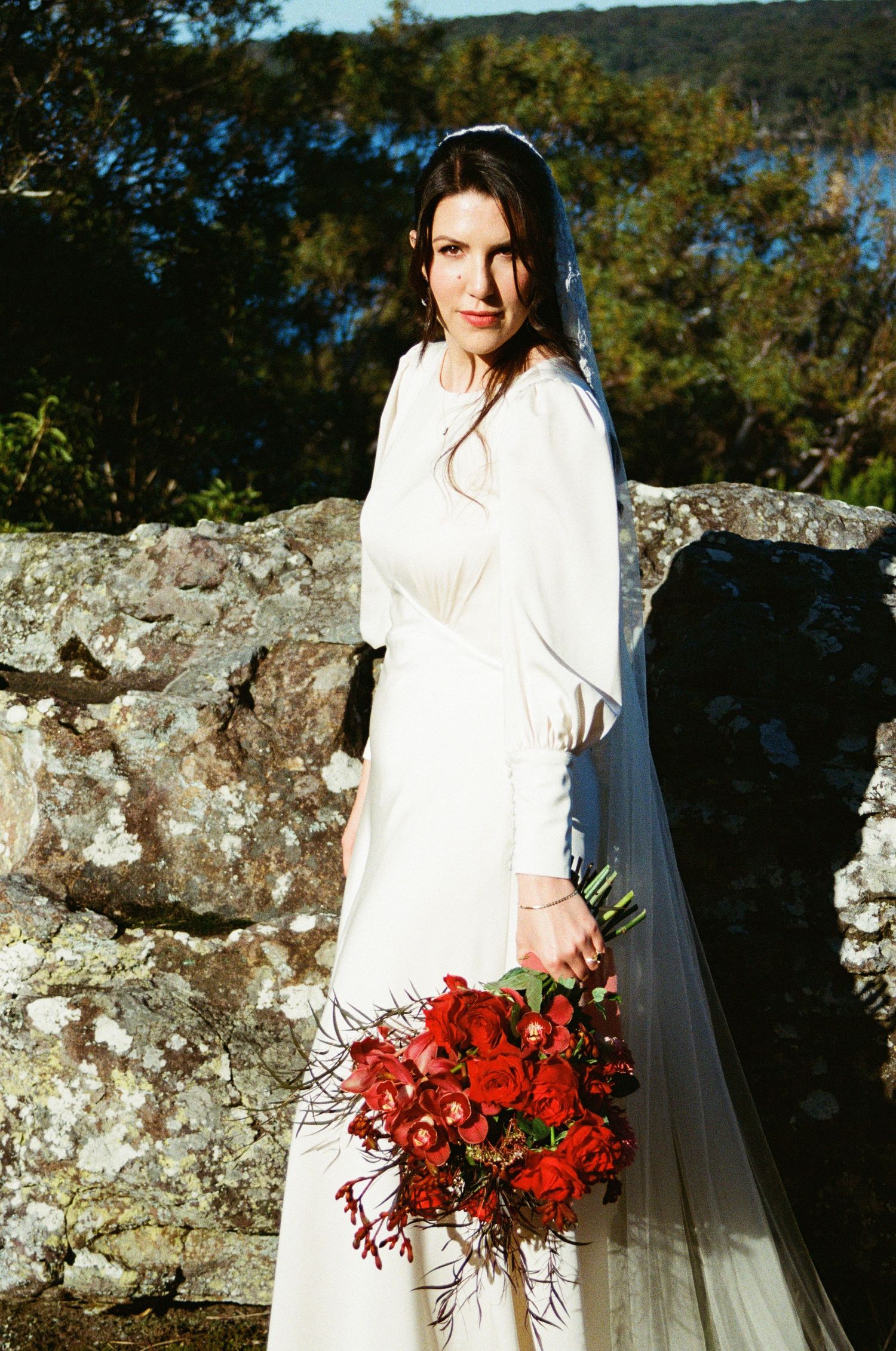 A bride in a long-sleeved white dress poses against stone wall holding vibrant red bouquet.