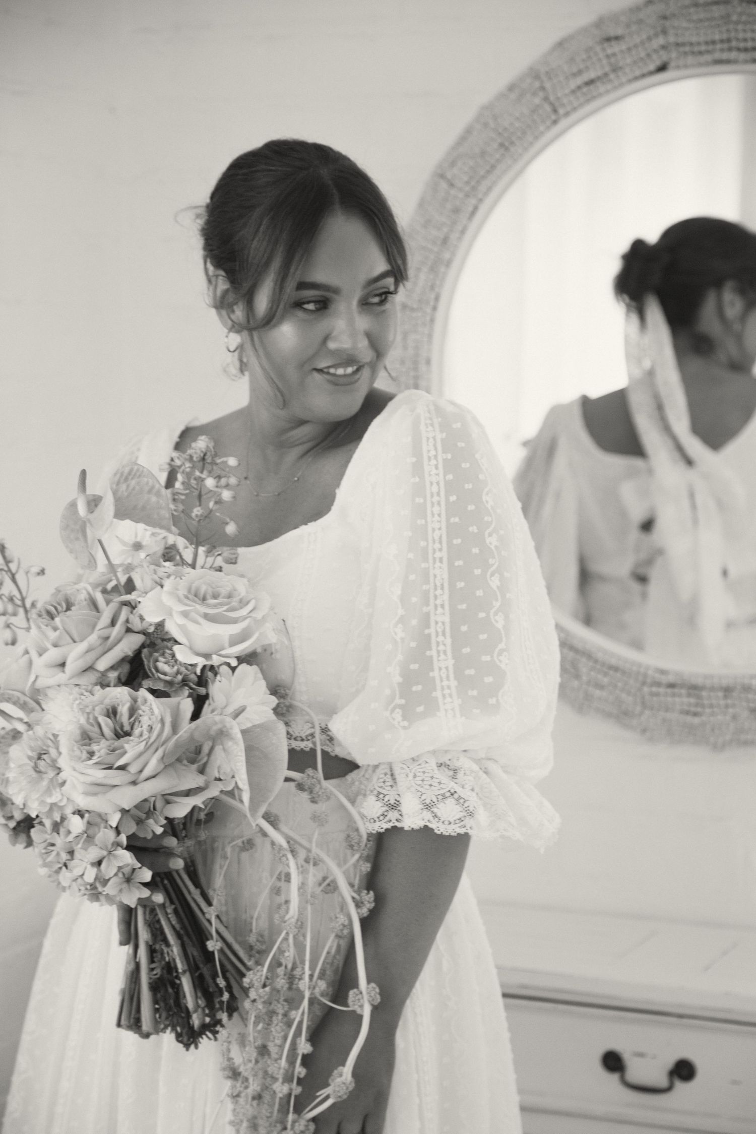 A bride in a vintage-inspired lace dress holds a bouquet while standing near an ornate mirror.