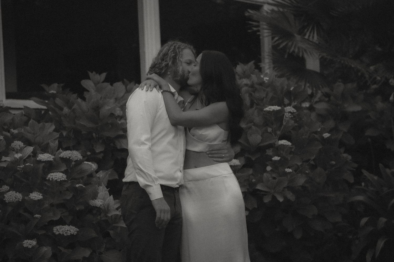 A black and white romantic photograph of an intimate moment between a couple in a garden setting.