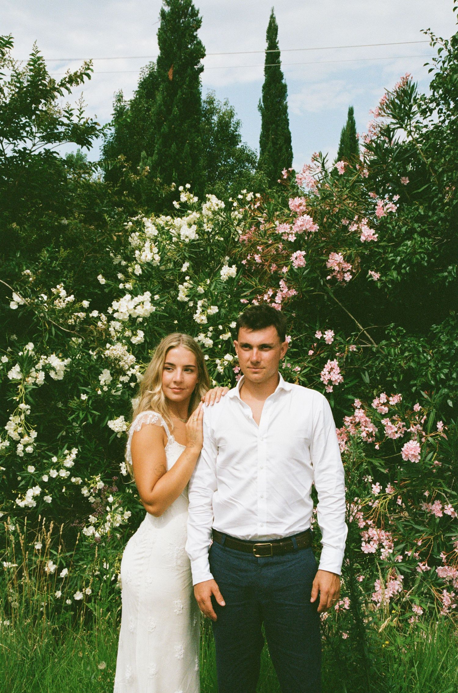A couple poses in a garden setting with blooming roses and cypress trees in the background.