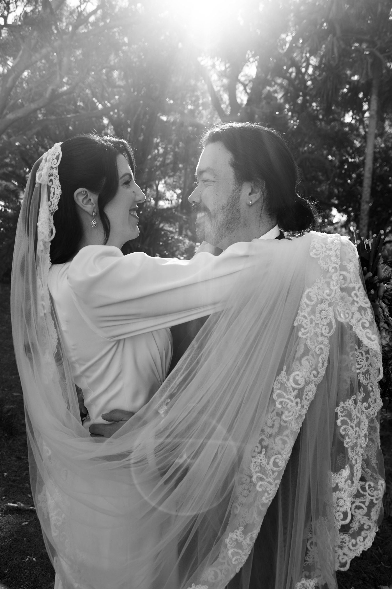 A black and white wedding portrait of a couple embracing outdoors with sunlight streaming through trees.