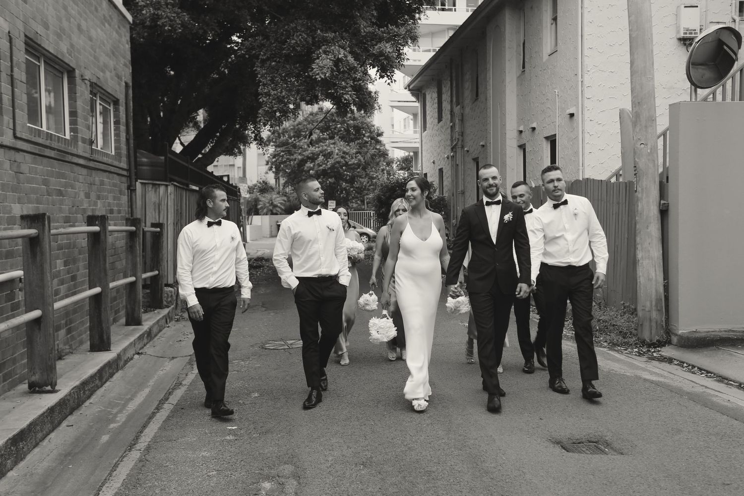 A black and white photograph shows a wedding party walking down a quiet city street together.