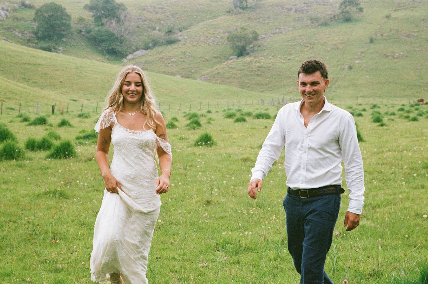Two people walk hand in hand along a winding country path with lush green hillsides in the background.