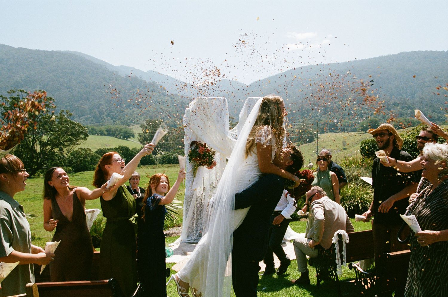 Wedding celebration with guests throwing flower petals outdoors against a mountain backdrop.