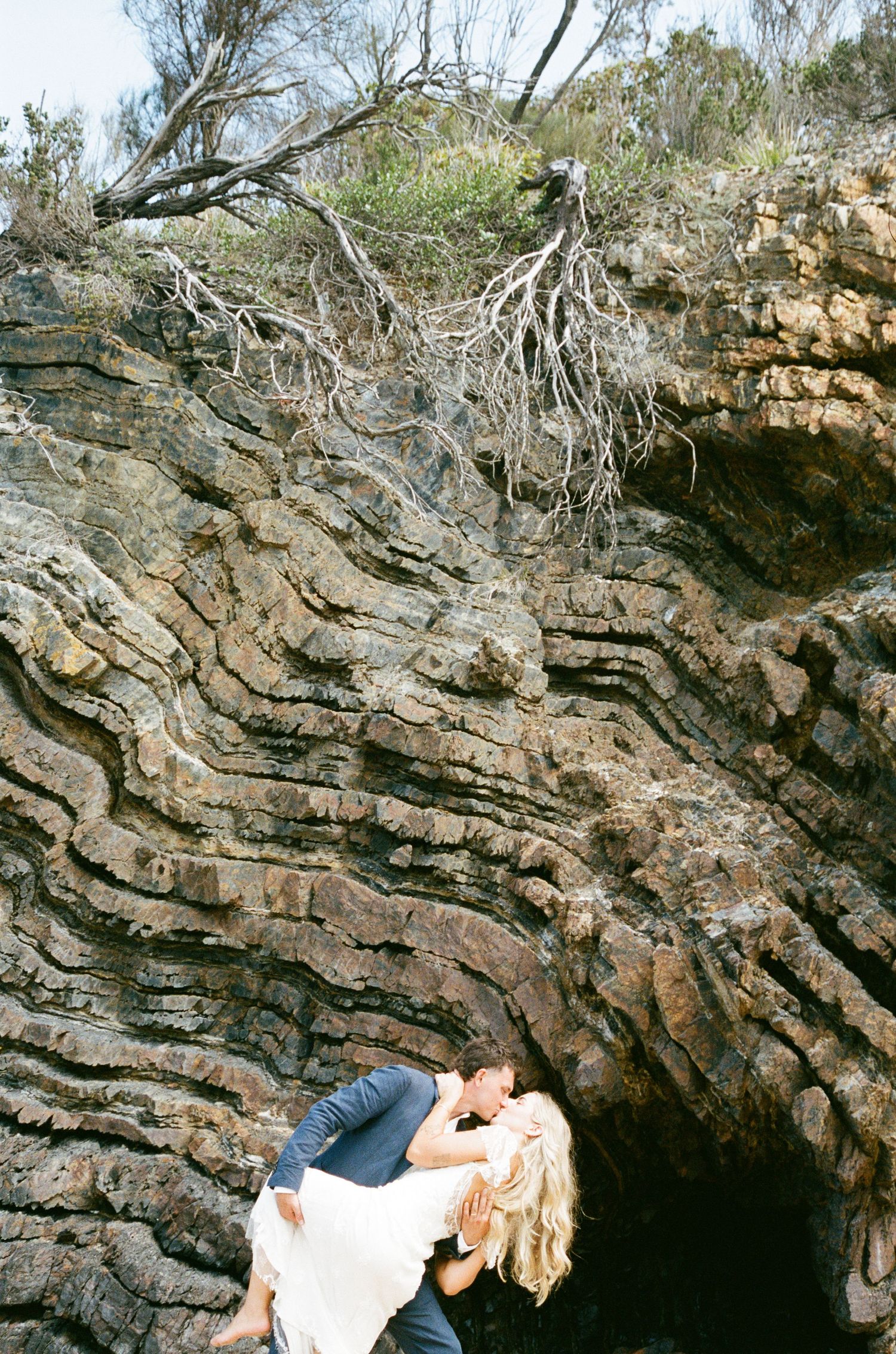 Two people relax under dramatic rock formations with twisted tree branches overhead.