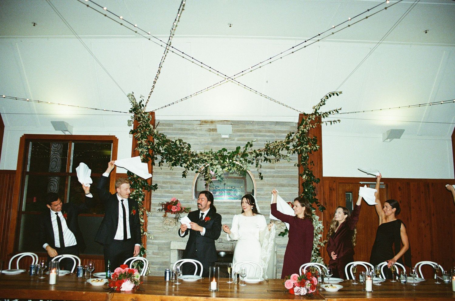 Wedding reception guests seated at a long table decorated with garlands celebrate with raised glasses in a wood-paneled room.