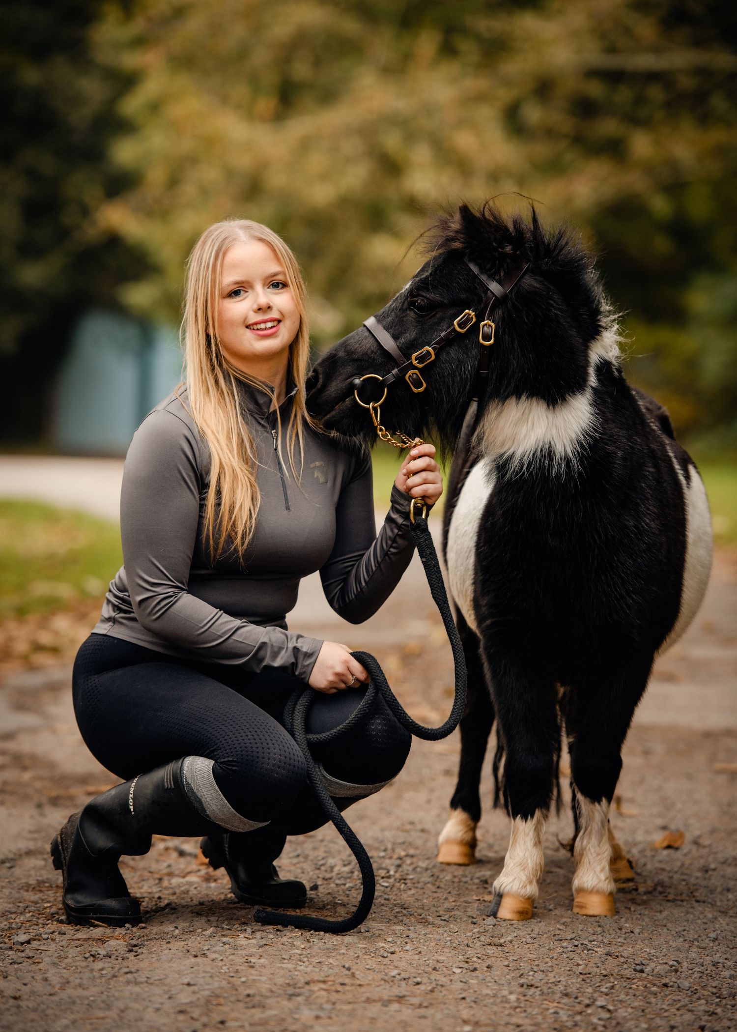 Young blonde woman in equestrian attire crouching beside a small black and white pony on a gravel path.