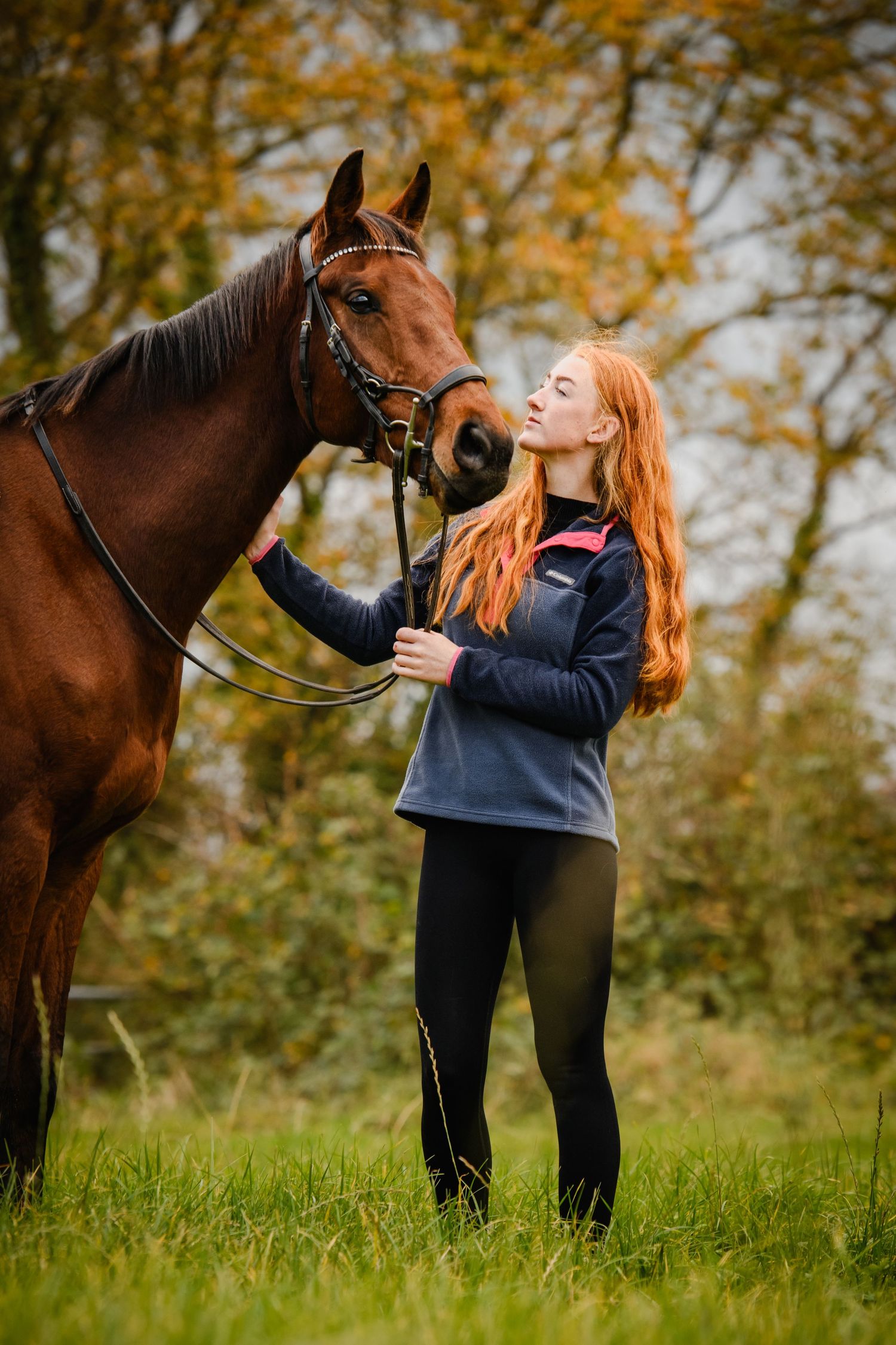 Red-haired girl standing peacefully beside a tall bay horse in autumn.