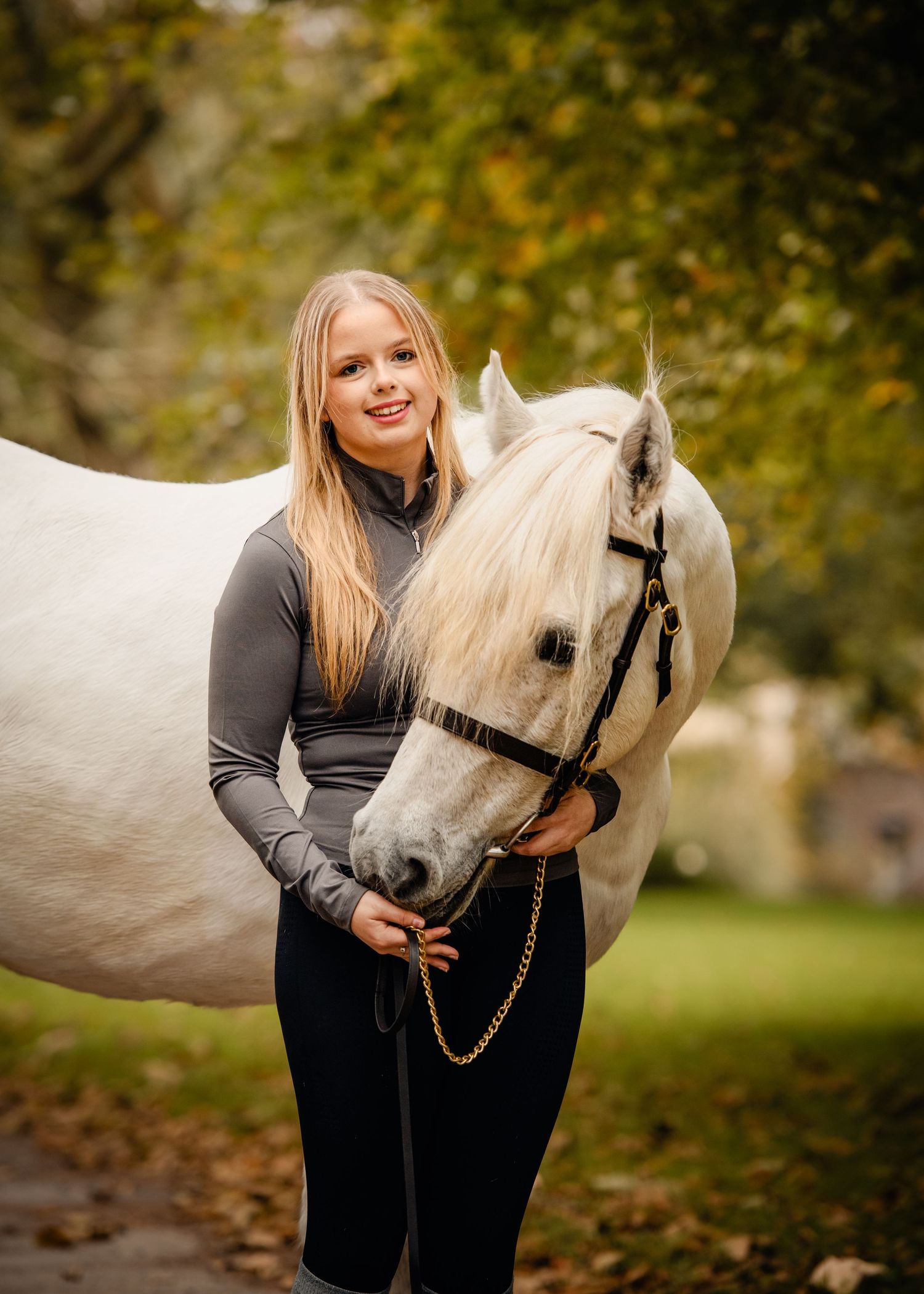 Young blonde girl smiling while embracing a white horse among autumn trees.