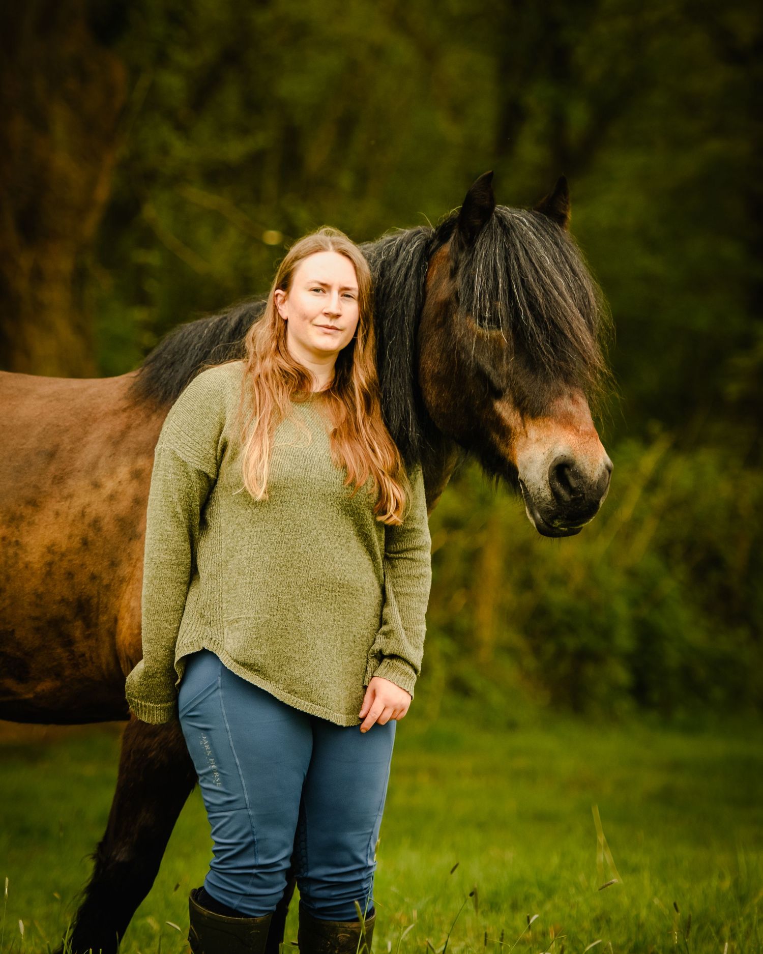 Young woman with long hair in green sweater stands beside a brown horse in a lush green field.