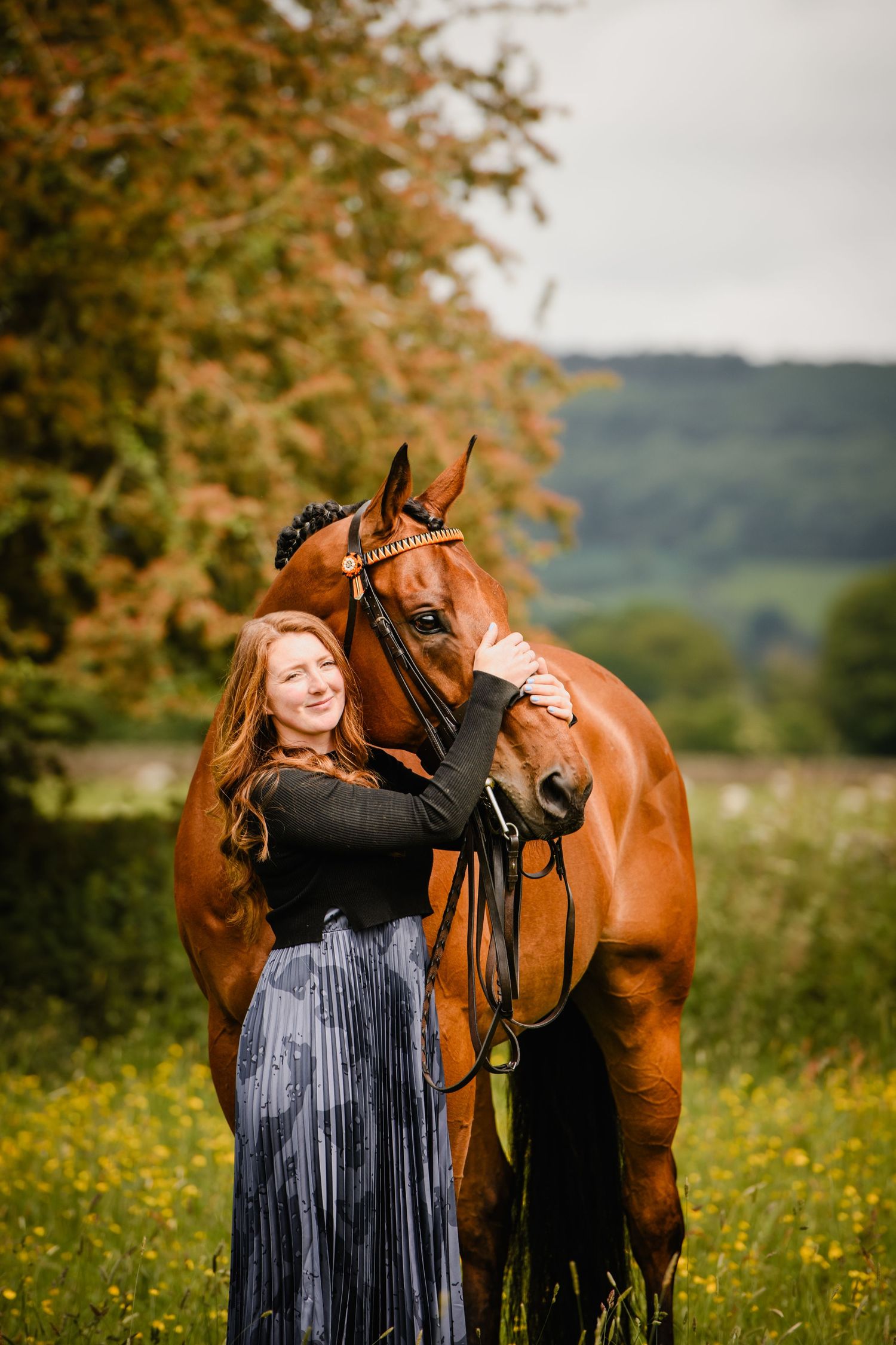A smiling redhead woman in a blue skirt embraces a chestnut horse in a lush green meadow.