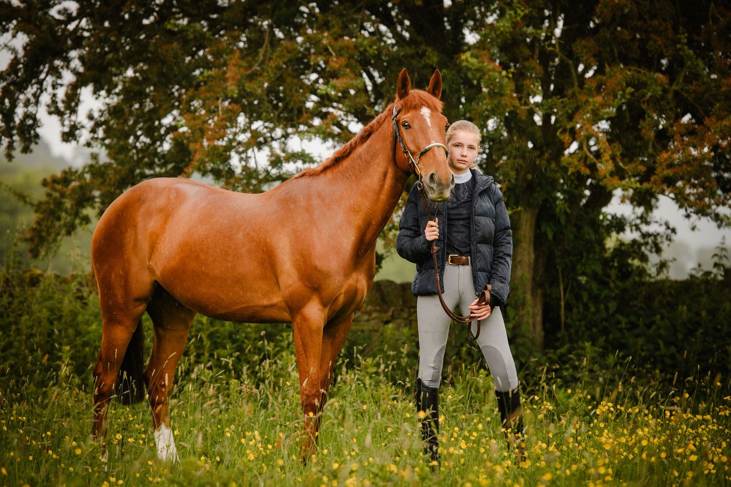 A blonde woman in equestrian attire stands beside a chestnut horse in a lush green meadow with trees.